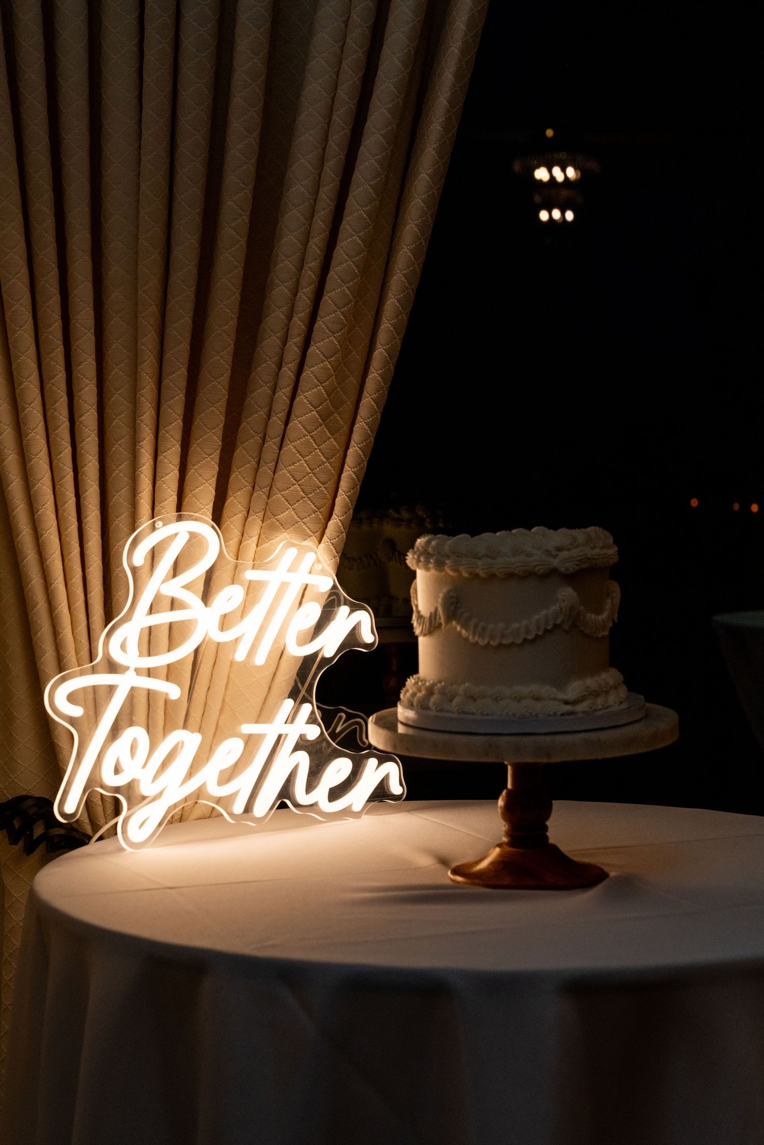 Wedding cake display with illuminated 'Better Together' neon sign in romantic lighting.