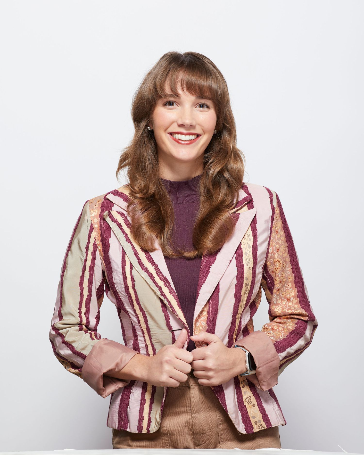 A person in a burgundy and beige striped blazer and purple top smiles warmly against a plain white background.