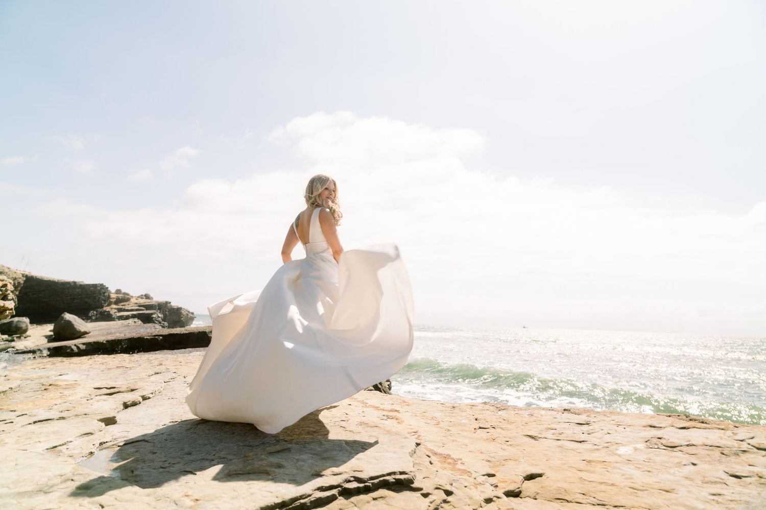 A flowing white wedding dress billows in the ocean breeze as someone stands on rocky coastal cliffs at sunset.