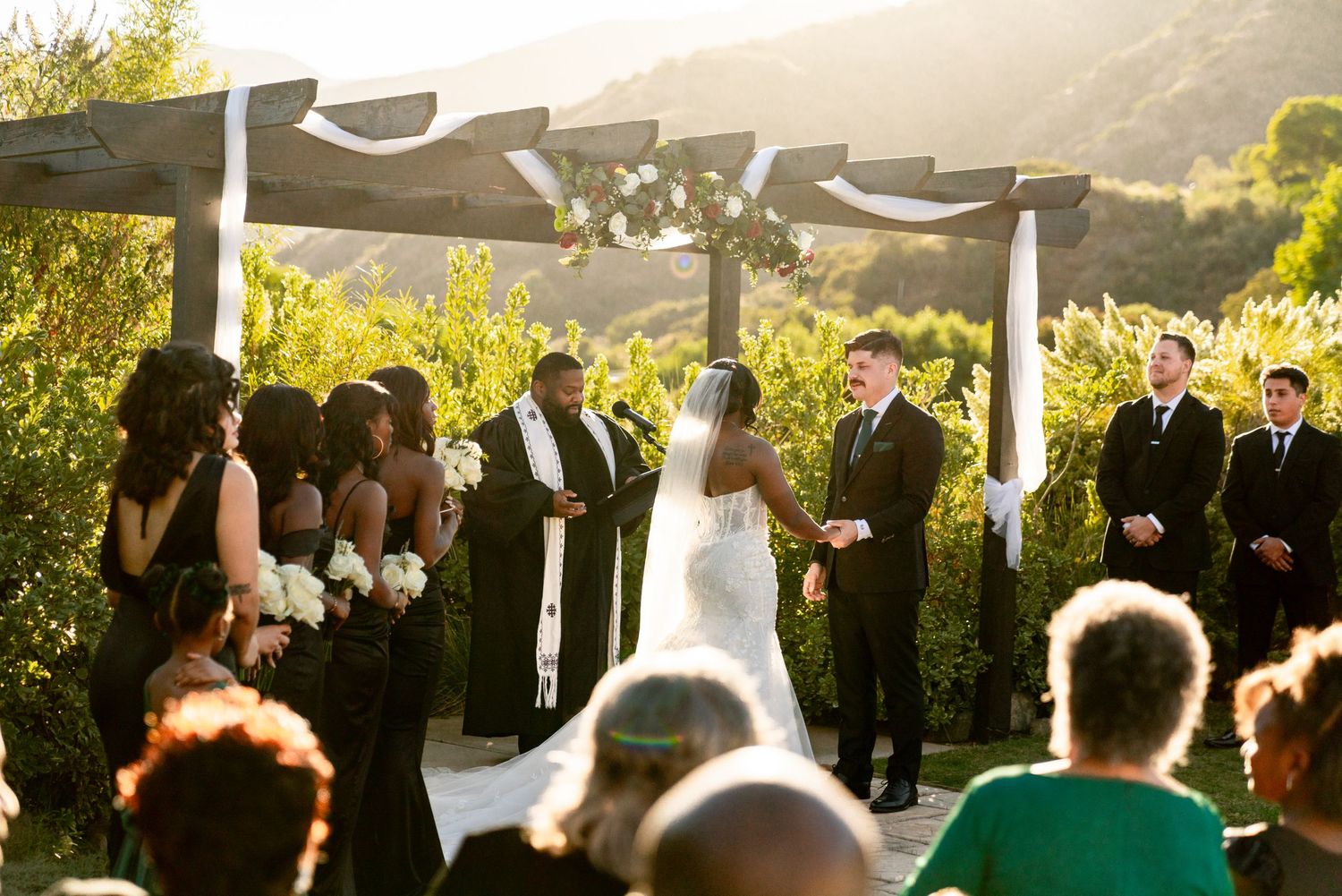Outdoor wedding ceremony under elegant draped pergola with mountain landscape at golden hour.