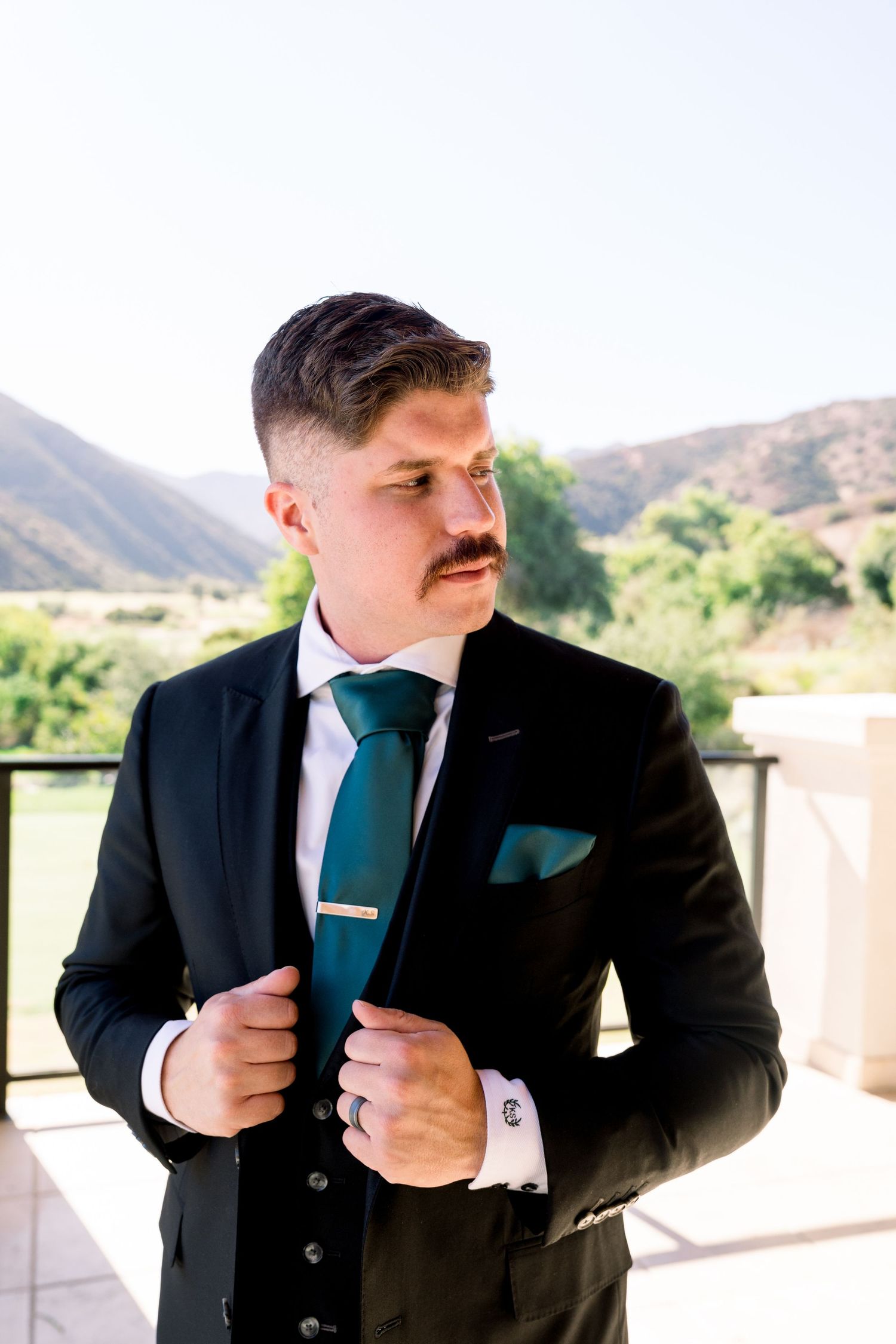 Person in formal black suit with teal tie adjusting jacket on outdoor balcony.