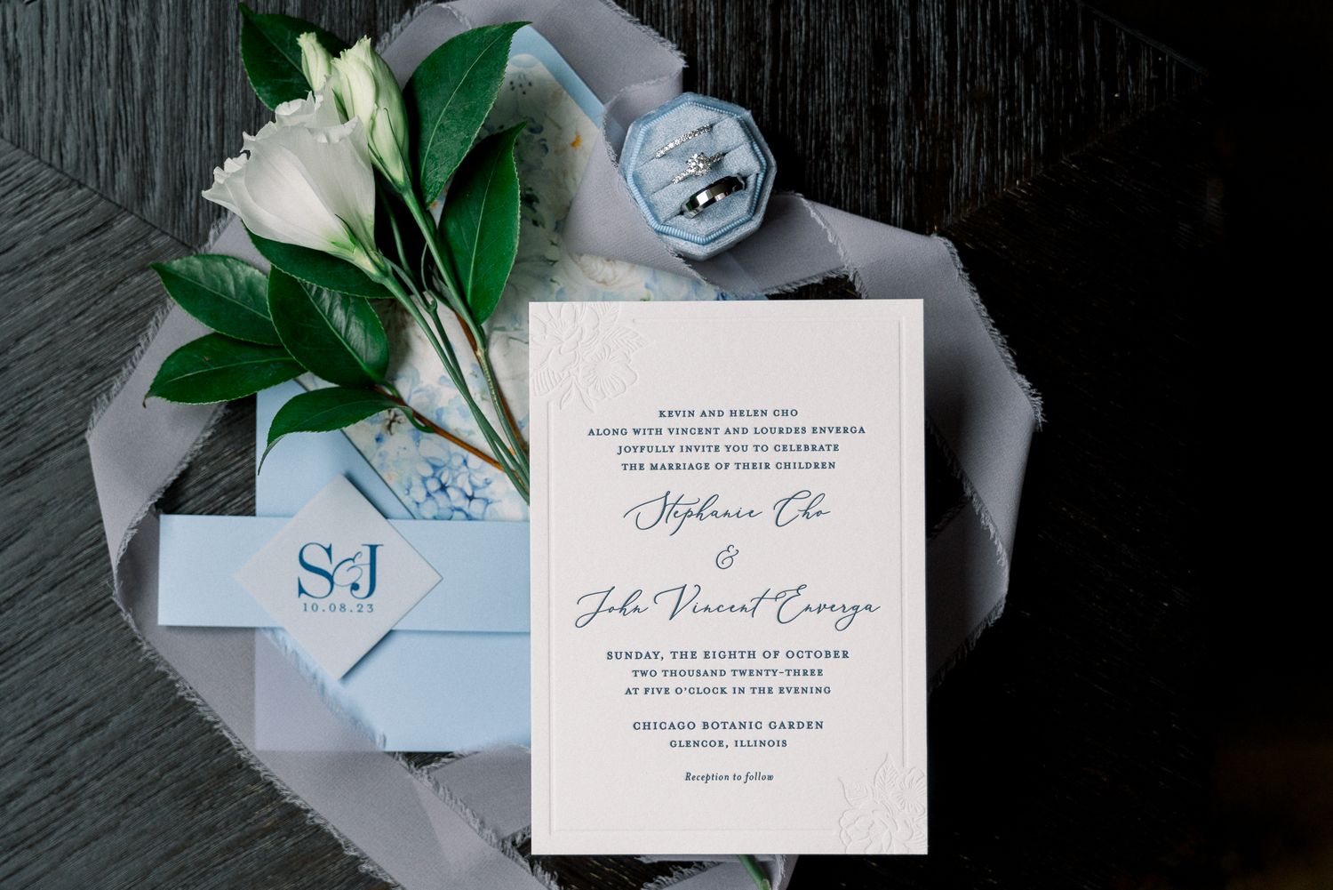 Elegant wedding invitation with white lily and blue details displayed on gray textured surface.