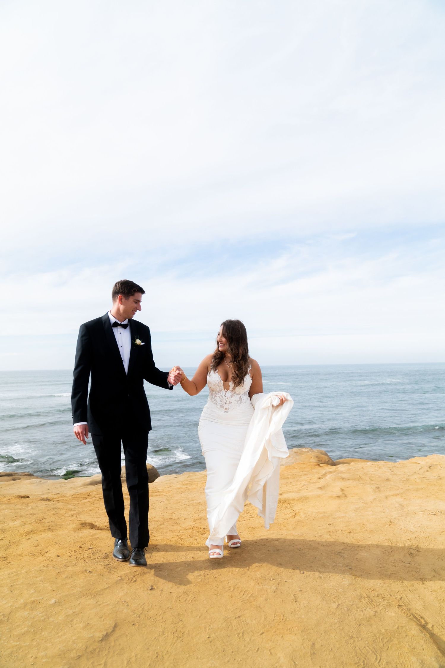 Newlyweds walk hand in hand along a scenic coastal cliff during their wedding photoshoot.
