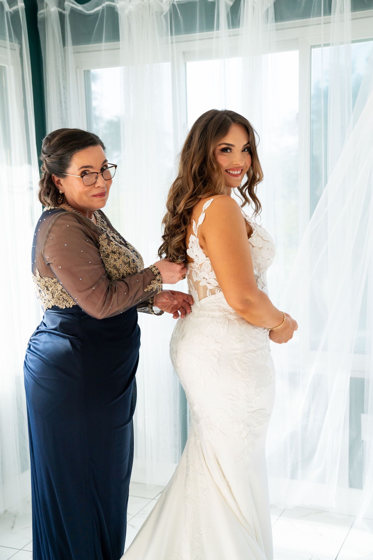 A bride in a fitted white lace dress gets help with final adjustments while standing near a window with sheer curtains.