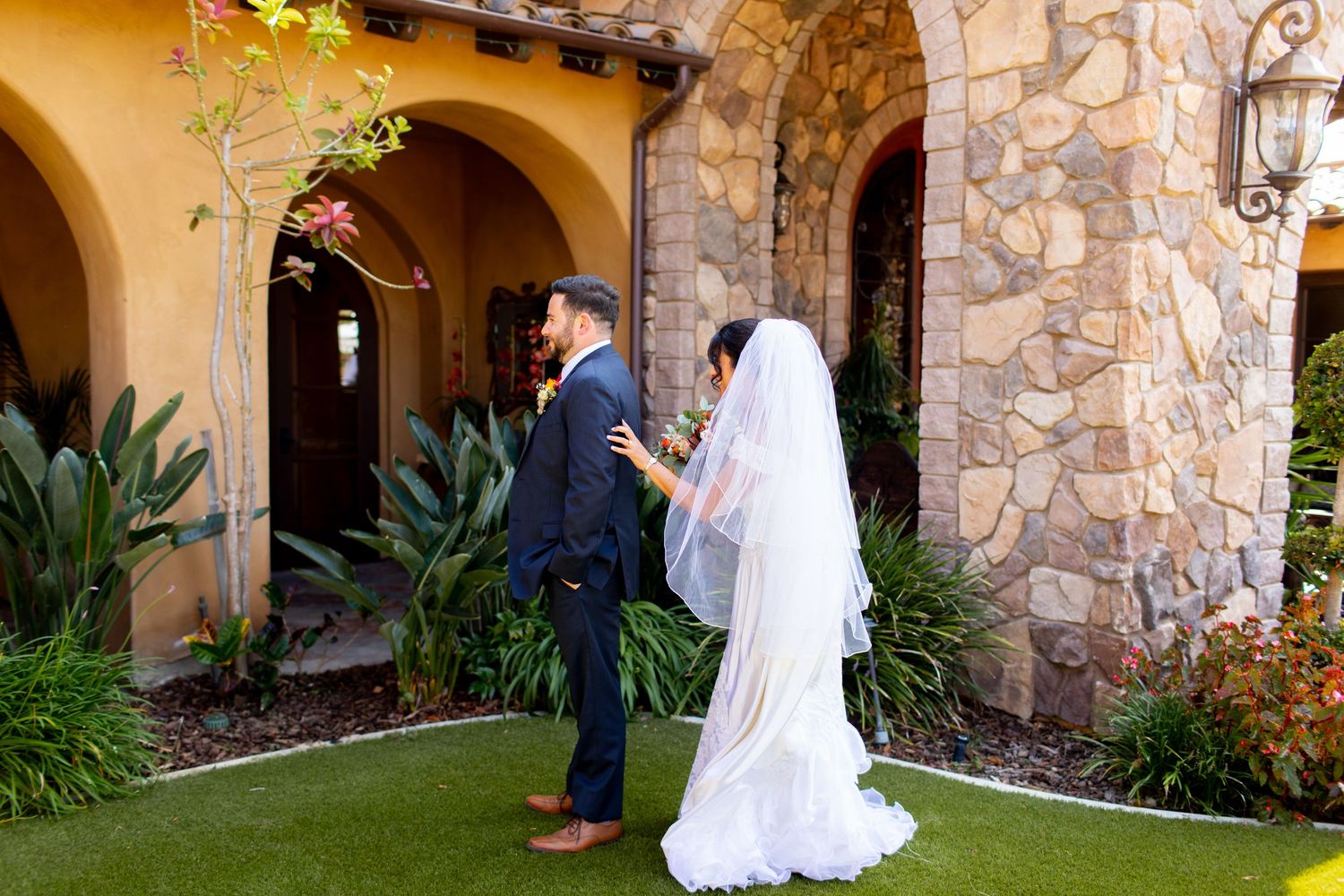 Wedding couple in formal attire standing in front of Mediterranean-style stone archway with lush garden.