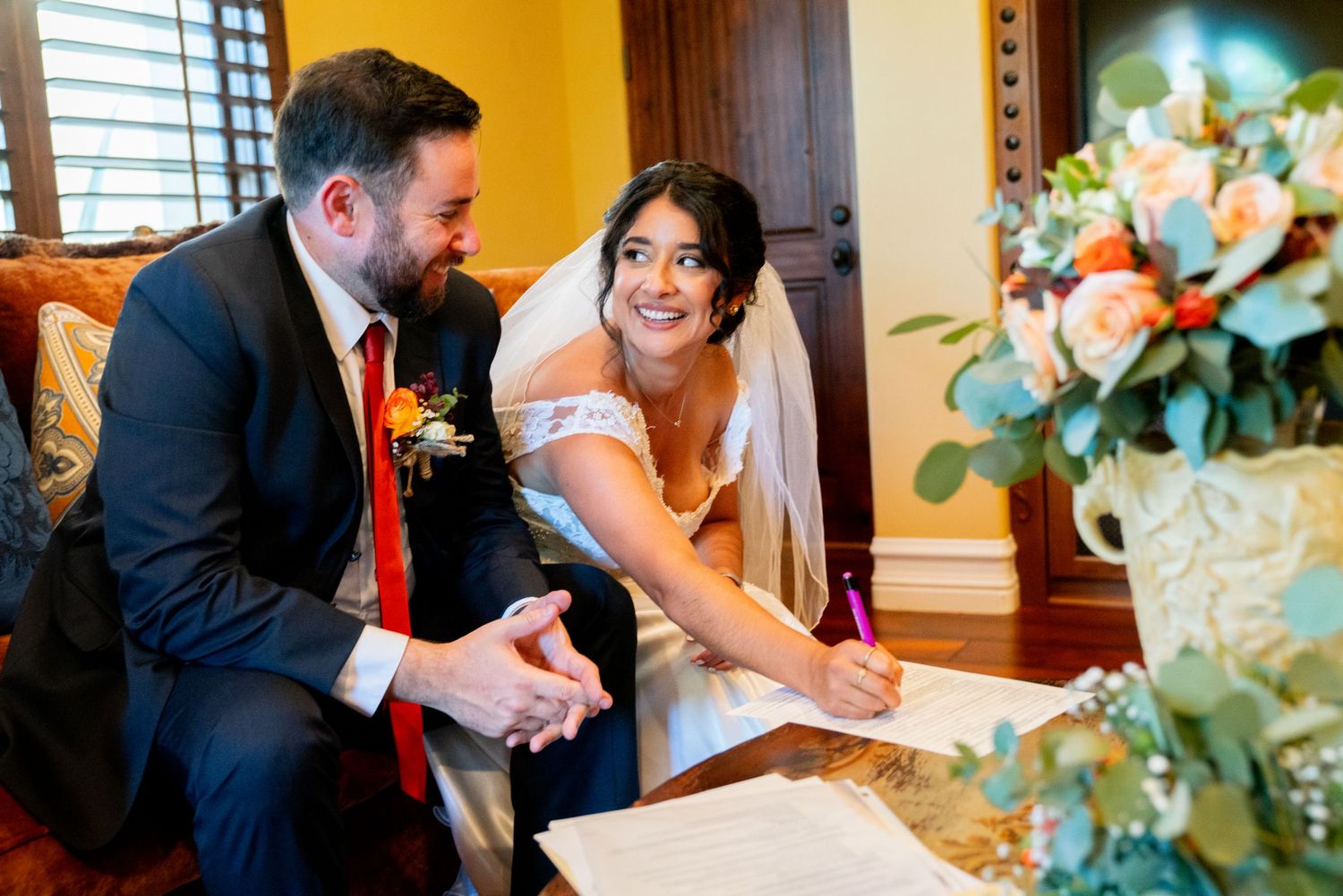 Couple signing their marriage license at wedding with floral decorations in background.