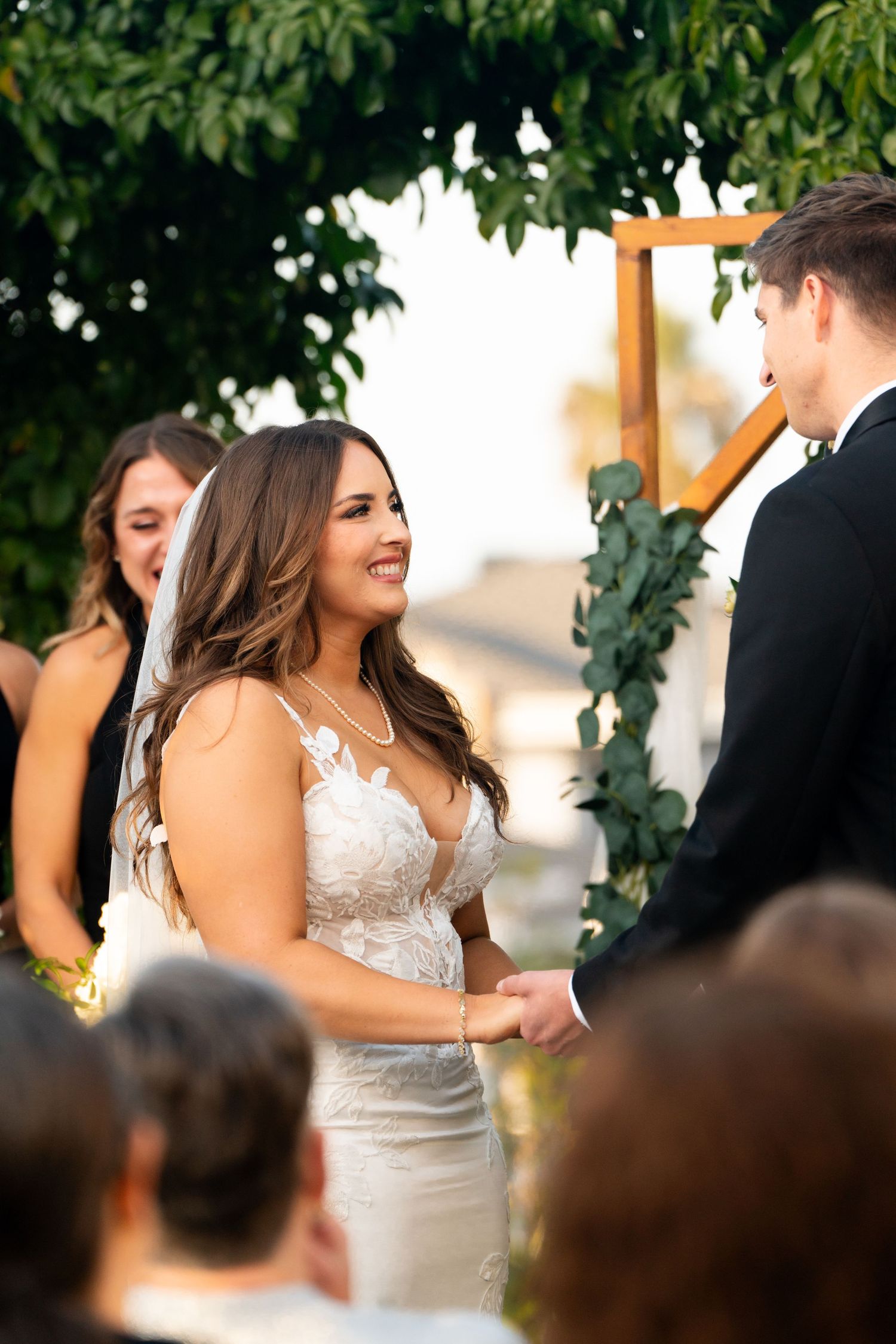A beautiful wedding ceremony takes place outdoors under a wooden arch decorated with greenery.