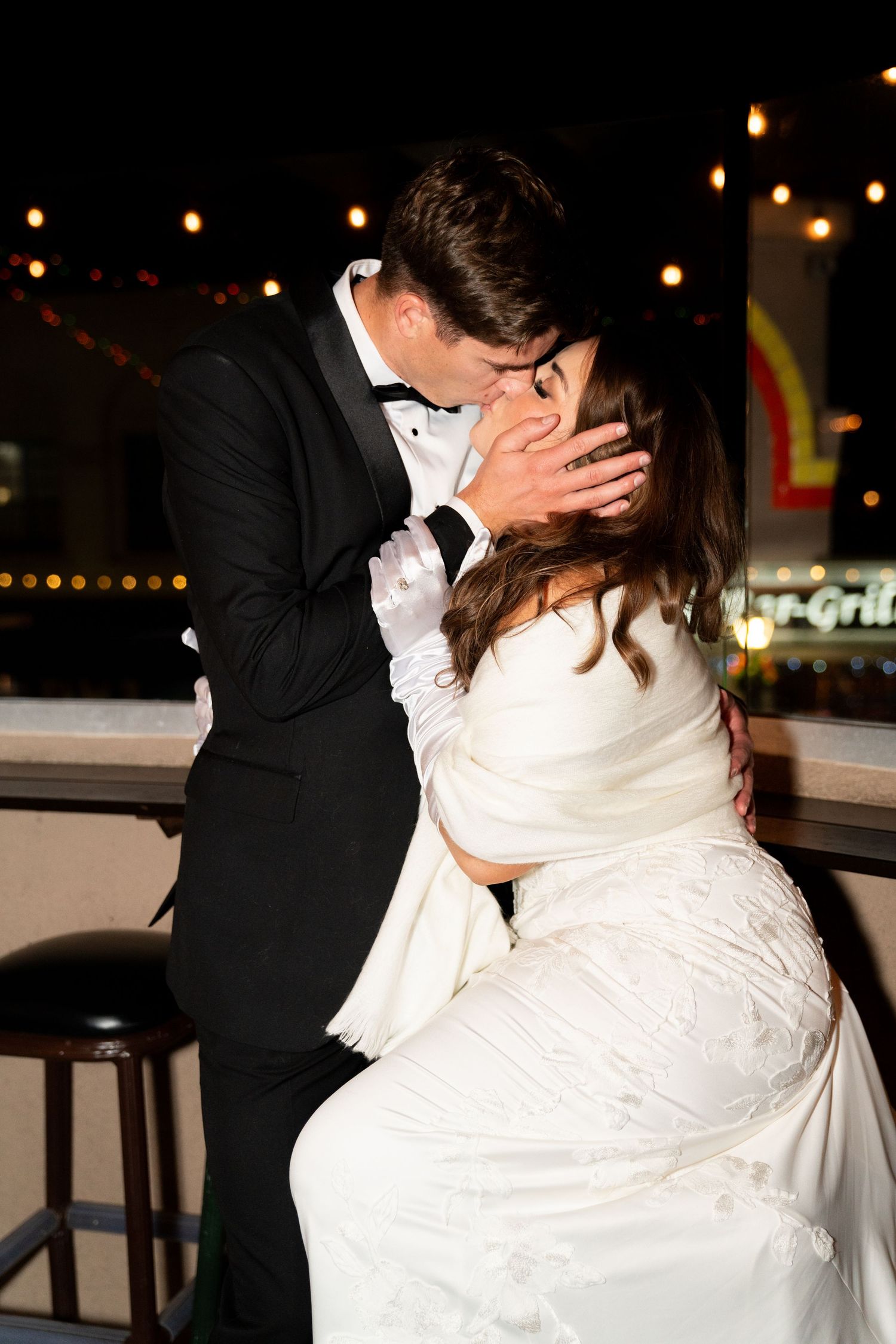 A couple shares a romantic kiss at their wedding reception with twinkling lights in the background.