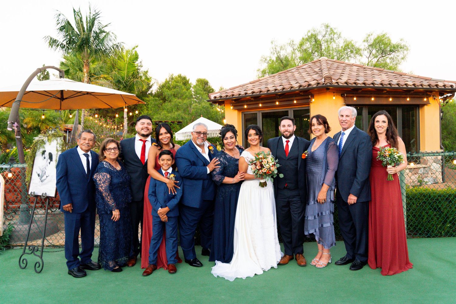 Wedding party gathered on green carpet in front of Spanish-style building with hanging lights and palm trees.