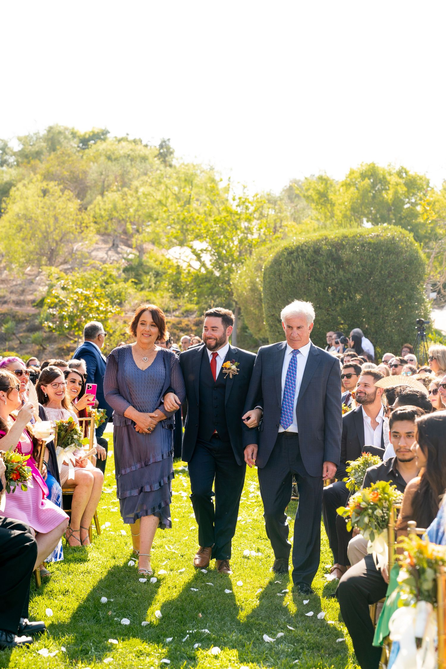 A sunlit outdoor wedding ceremony takes place in a garden setting with floral arrangements along the grassy aisle.