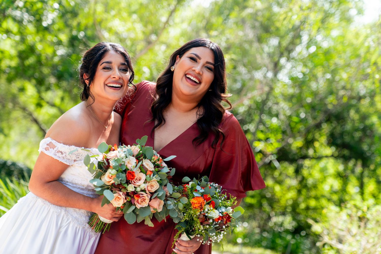 Two laughing people holding beautiful bouquets of peach roses and eucalyptus against a green outdoor background.