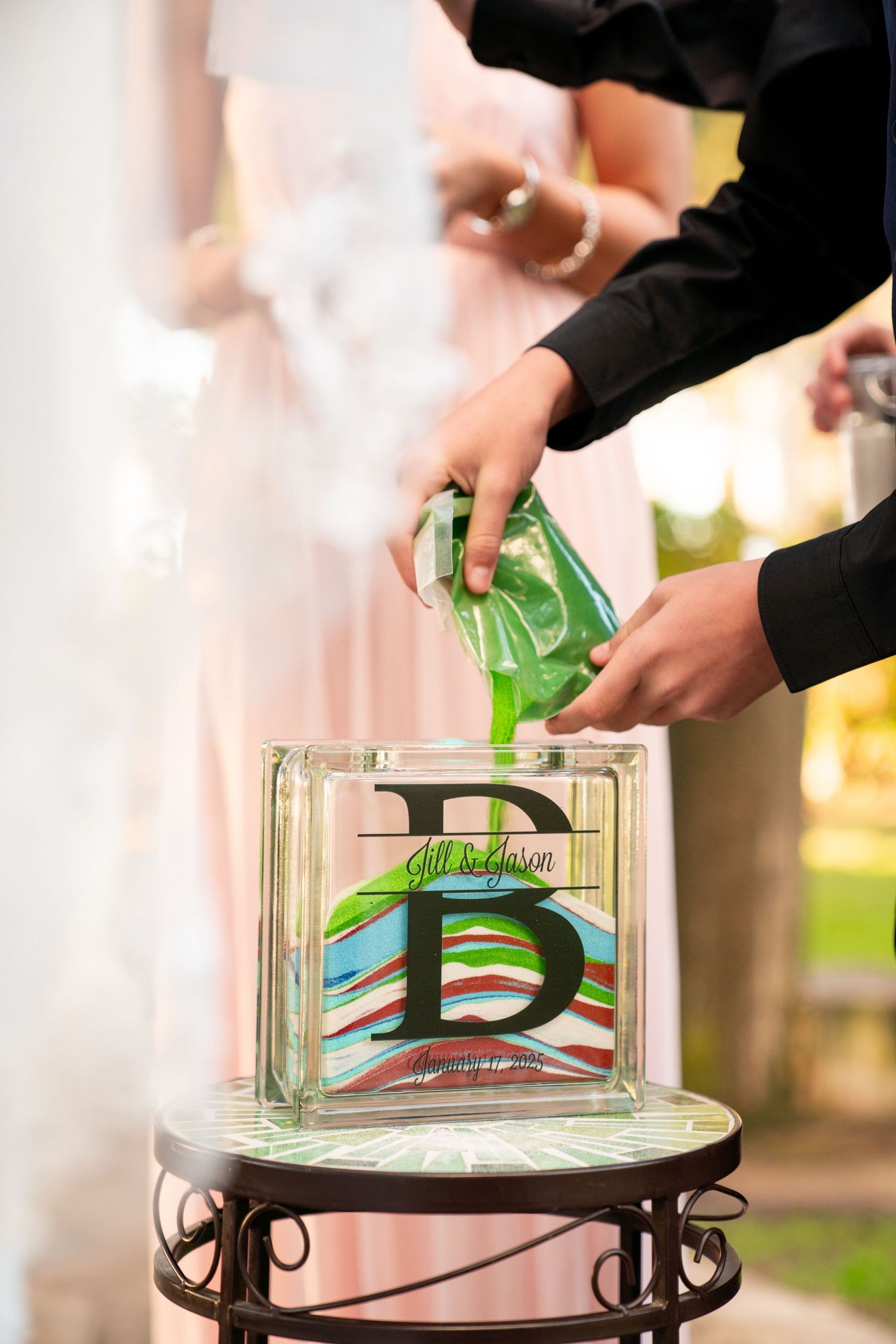 Hands pouring green sand into a decorative glass block during a unity sand ceremony at a wedding.