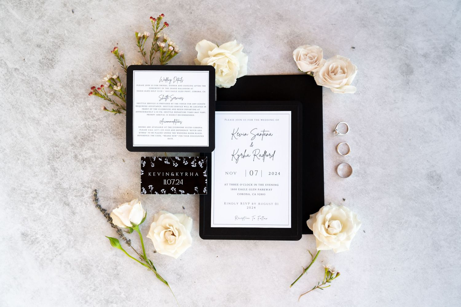 Wedding invitations in black and white styling with cream roses and rings on marble surface.