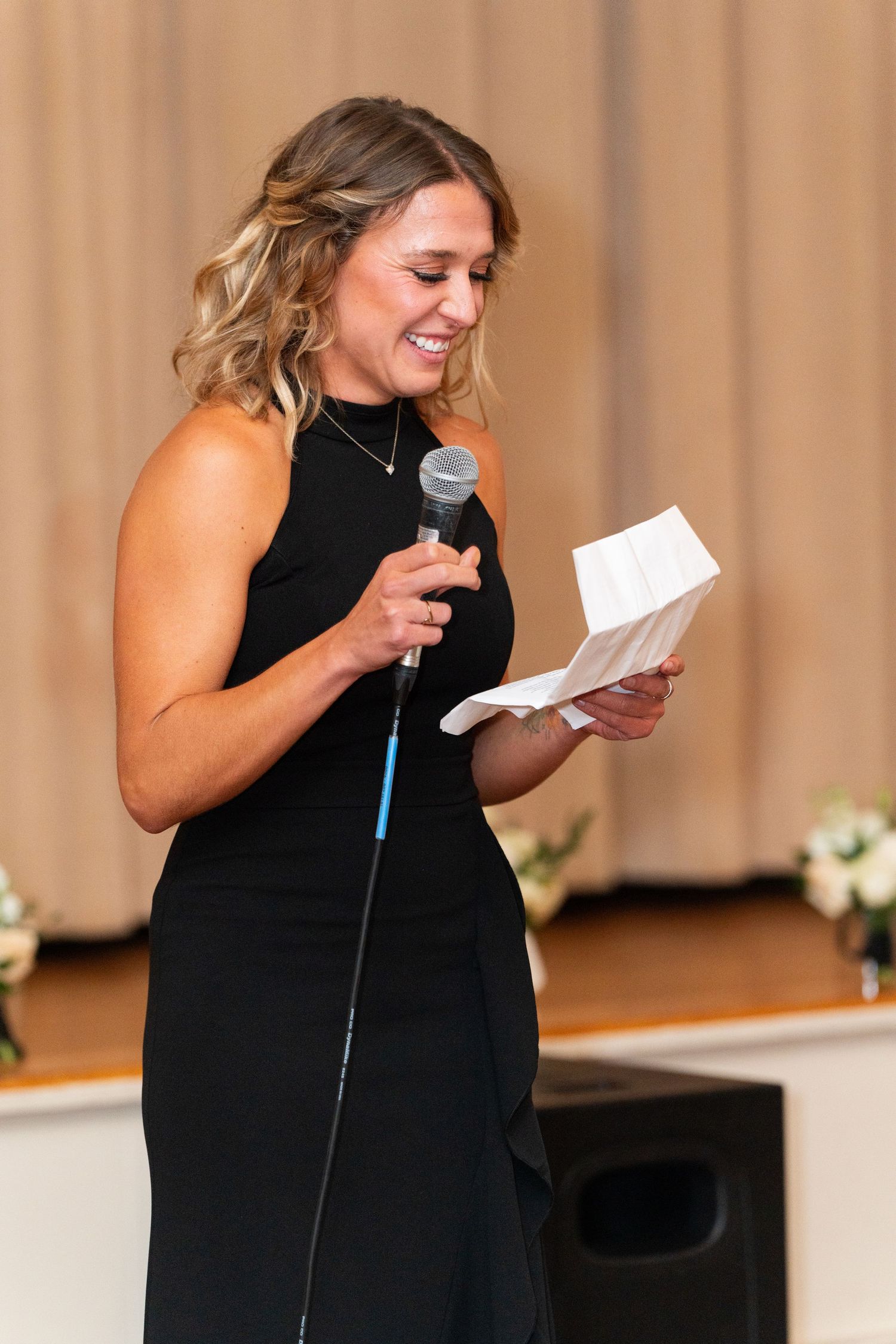 A woman in a black dress delivers a speech while holding a microphone and notes at a formal event.