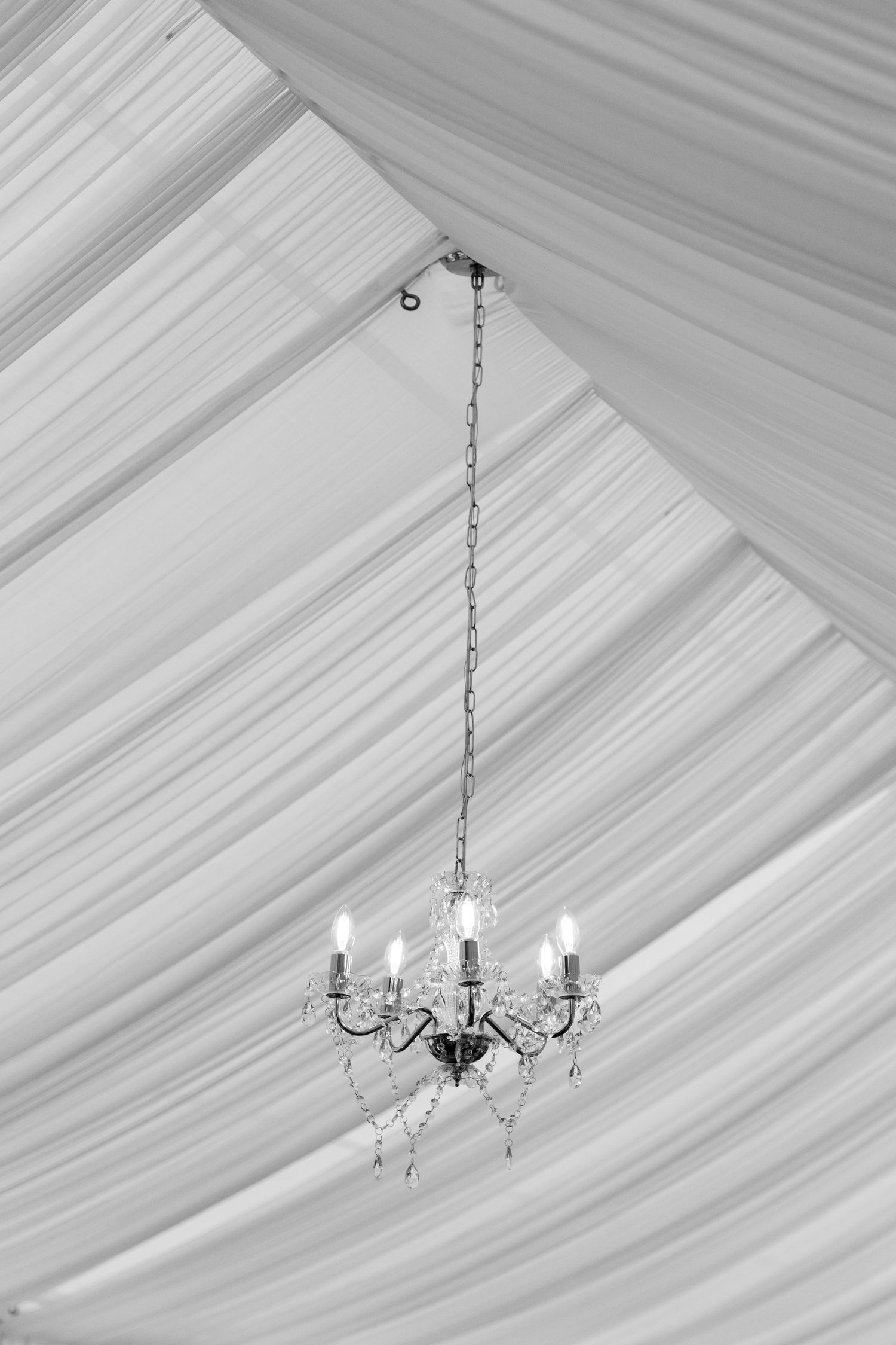 An elegant crystal chandelier hangs from draped white fabric ceiling at an indoor wedding venue.