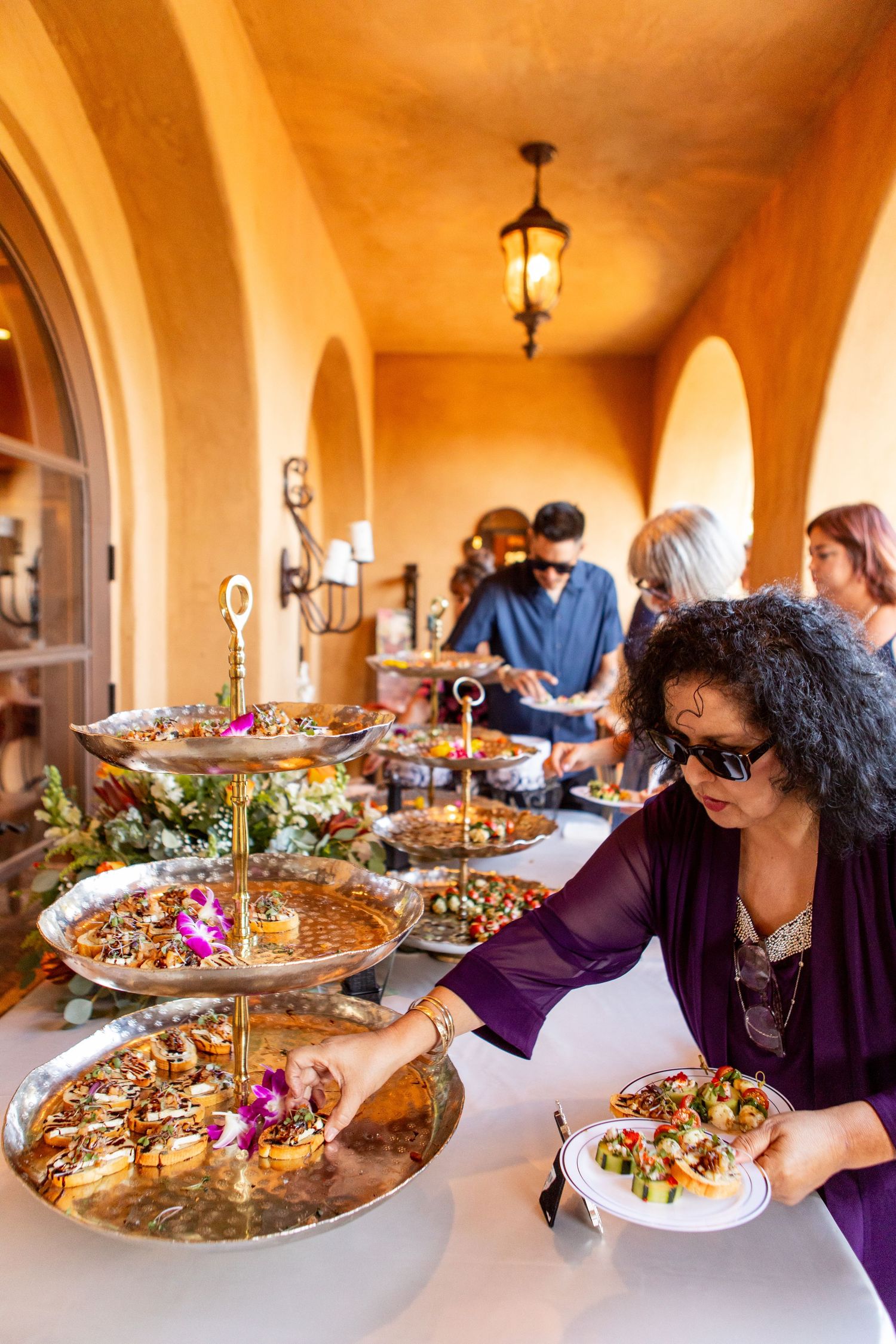 Wedding guests serve themselves from ornate tiered platters at a buffet under Mediterranean-style arches.