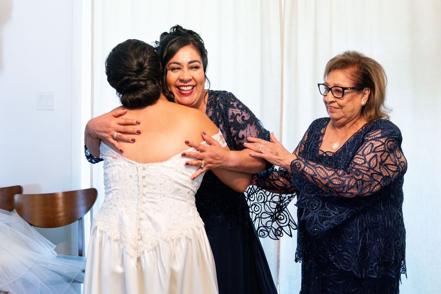 A bride in a white wedding dress shares an emotional moment with two women wearing navy lace dresses.