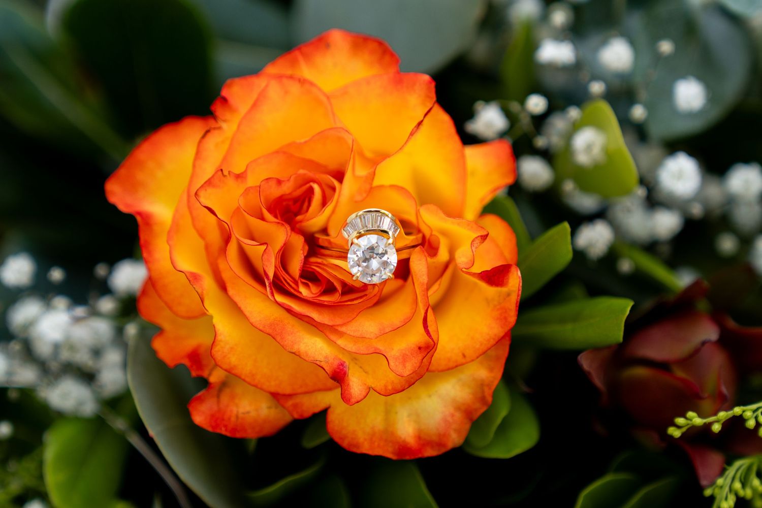 An elegant diamond ring nestled in the center of a vibrant orange rose with baby's breath flowers in the background.