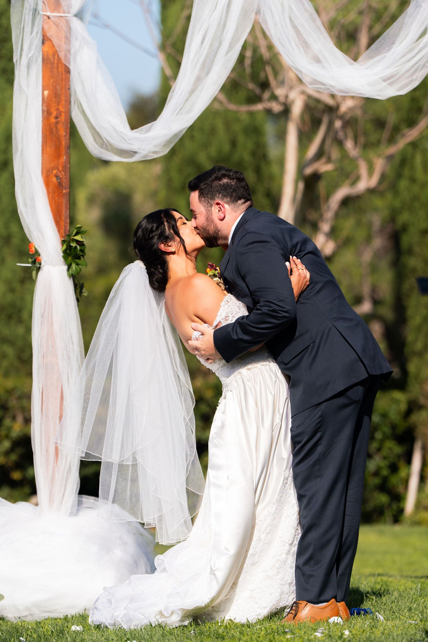 Romantic wedding kiss under white fabric draped archway in outdoor garden setting.