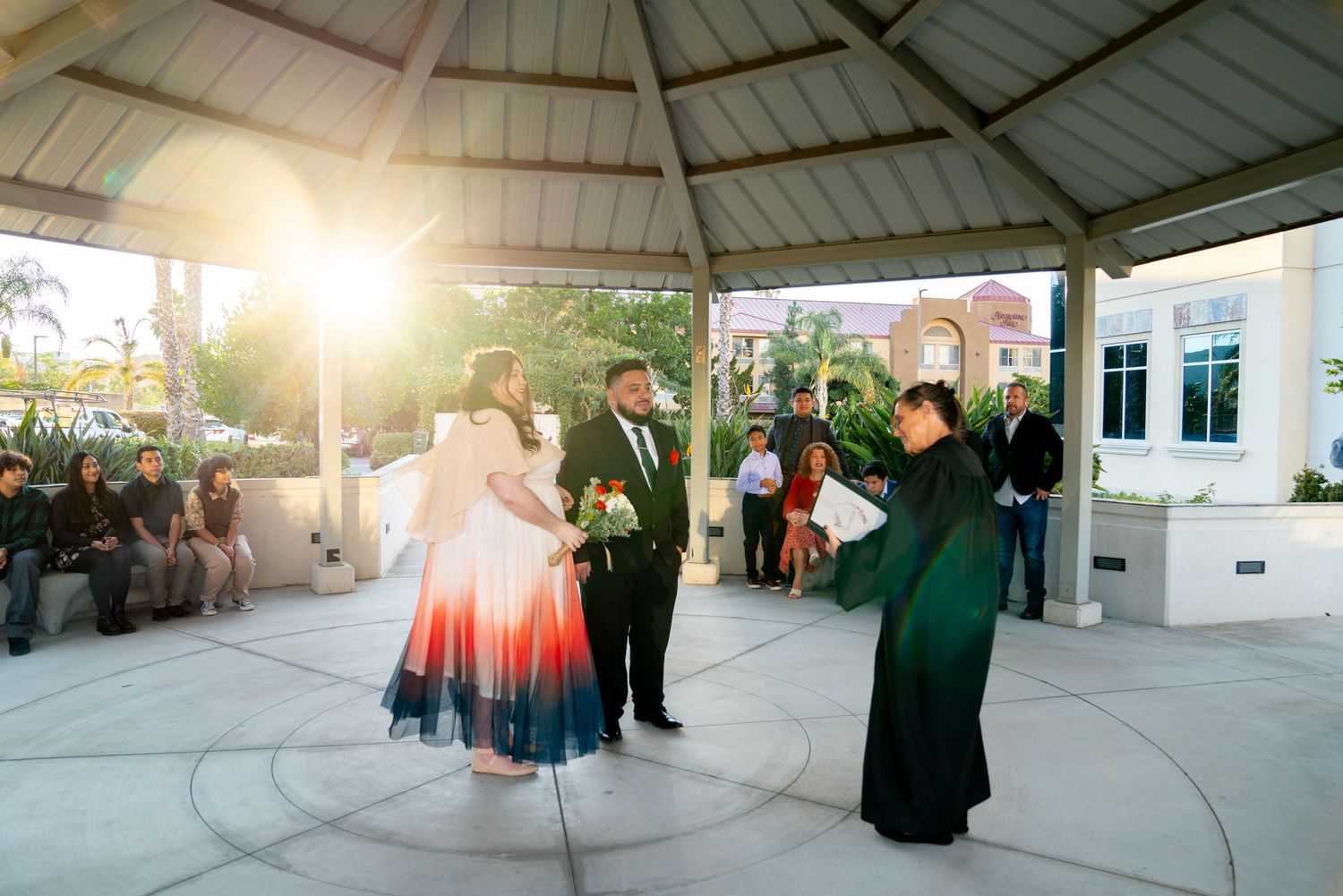 A sun-lit outdoor gazebo scene with formal attire-clad guests dancing at a celebration.