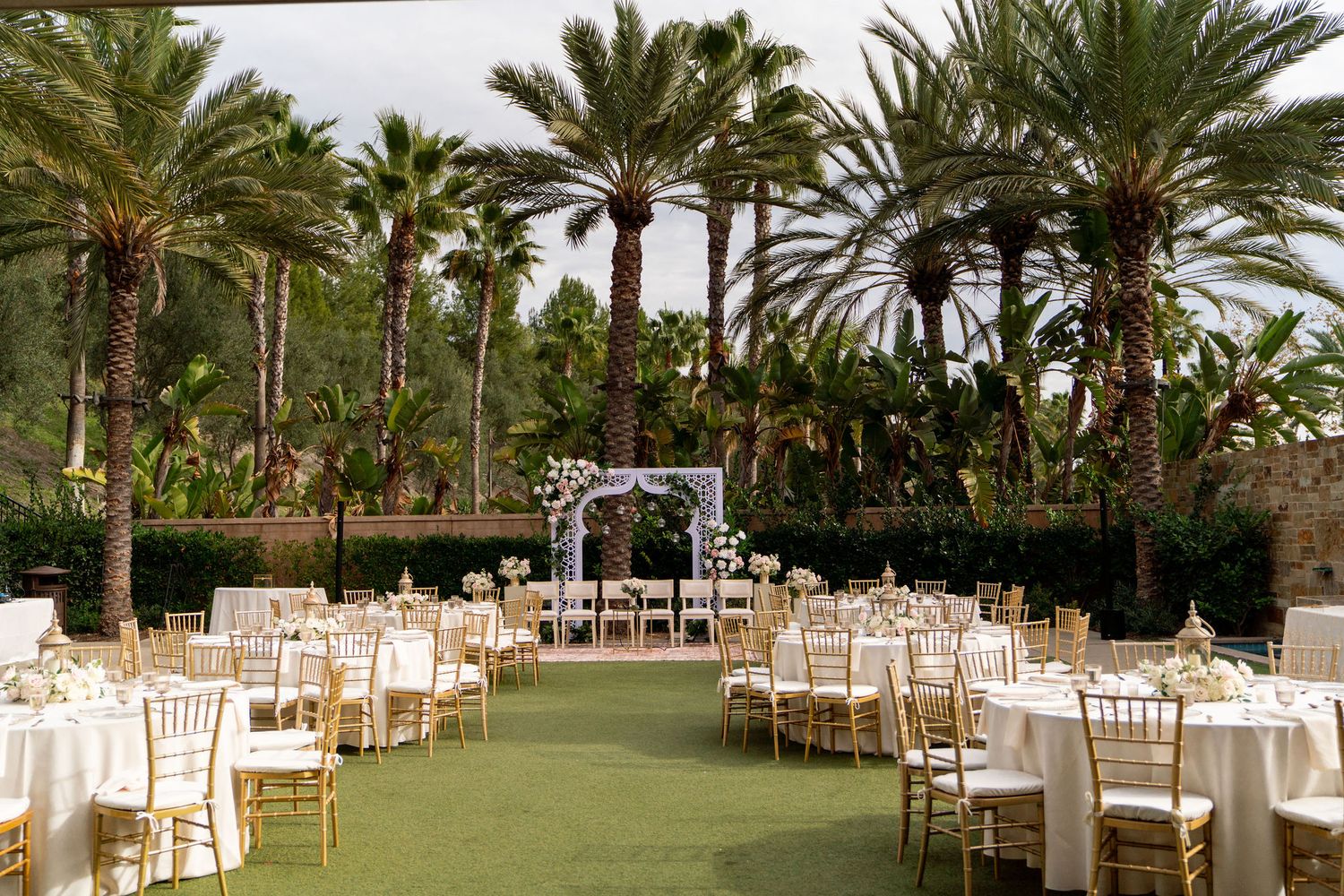 Elegant outdoor wedding venue setup with white chairs and tables arranged on grass lawn surrounded by tropical palm trees.