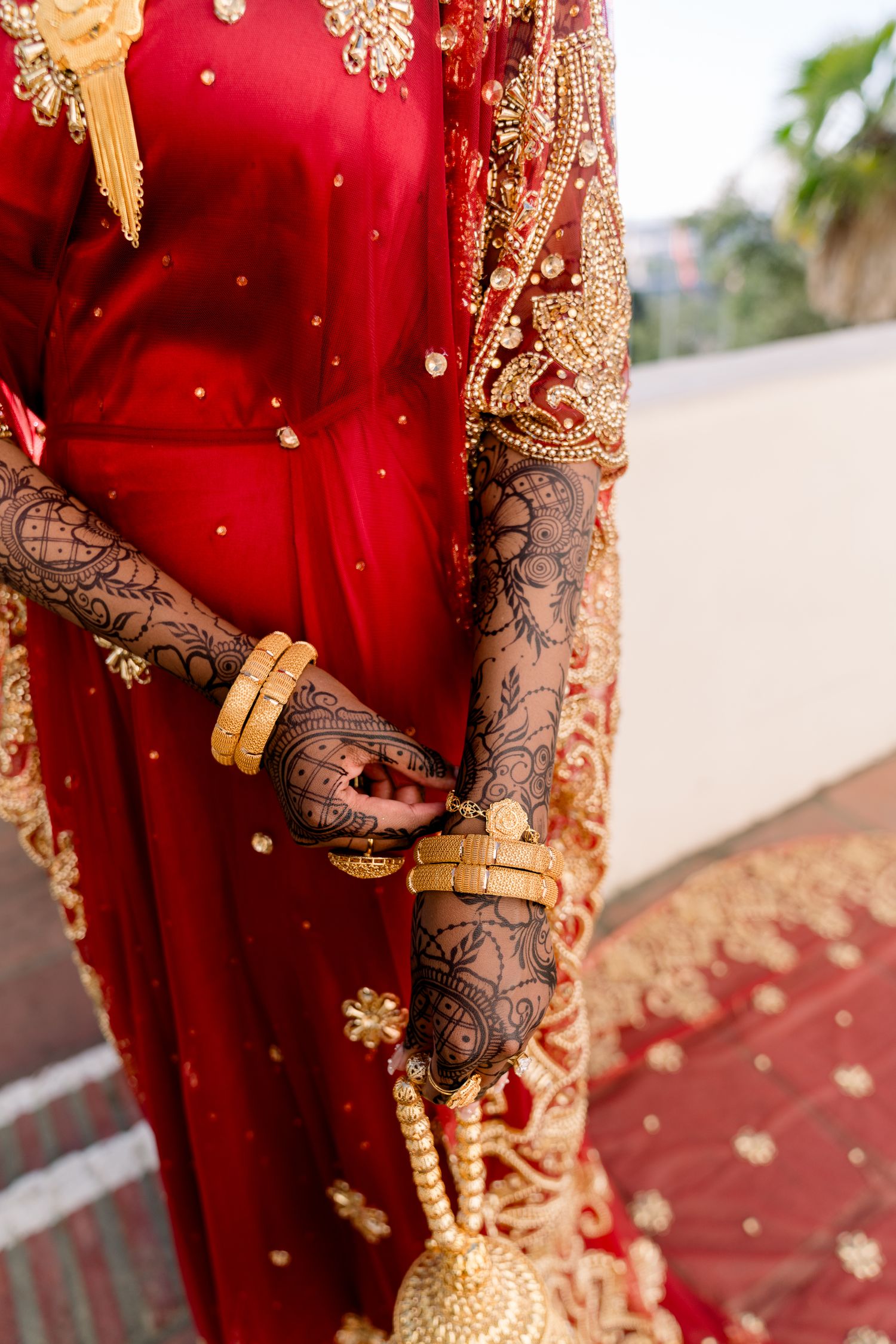 Beautiful henna-adorned hands wearing gold bangles against red bridal lehenga with golden embroidery and embellishments.