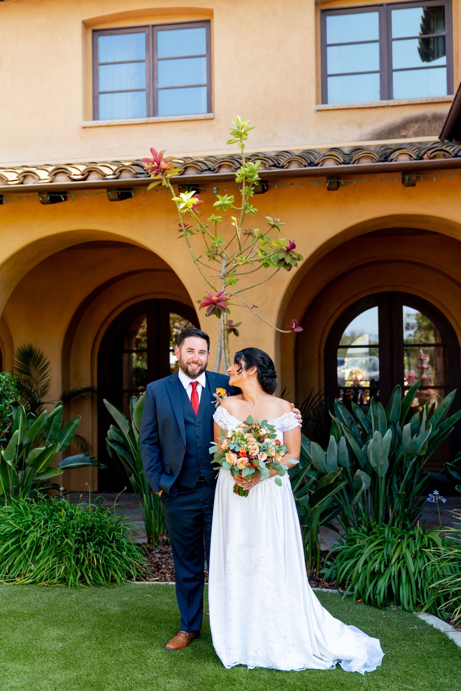 A wedding portrait in front of a Spanish colonial building with arched colonnade and tropical garden landscaping.