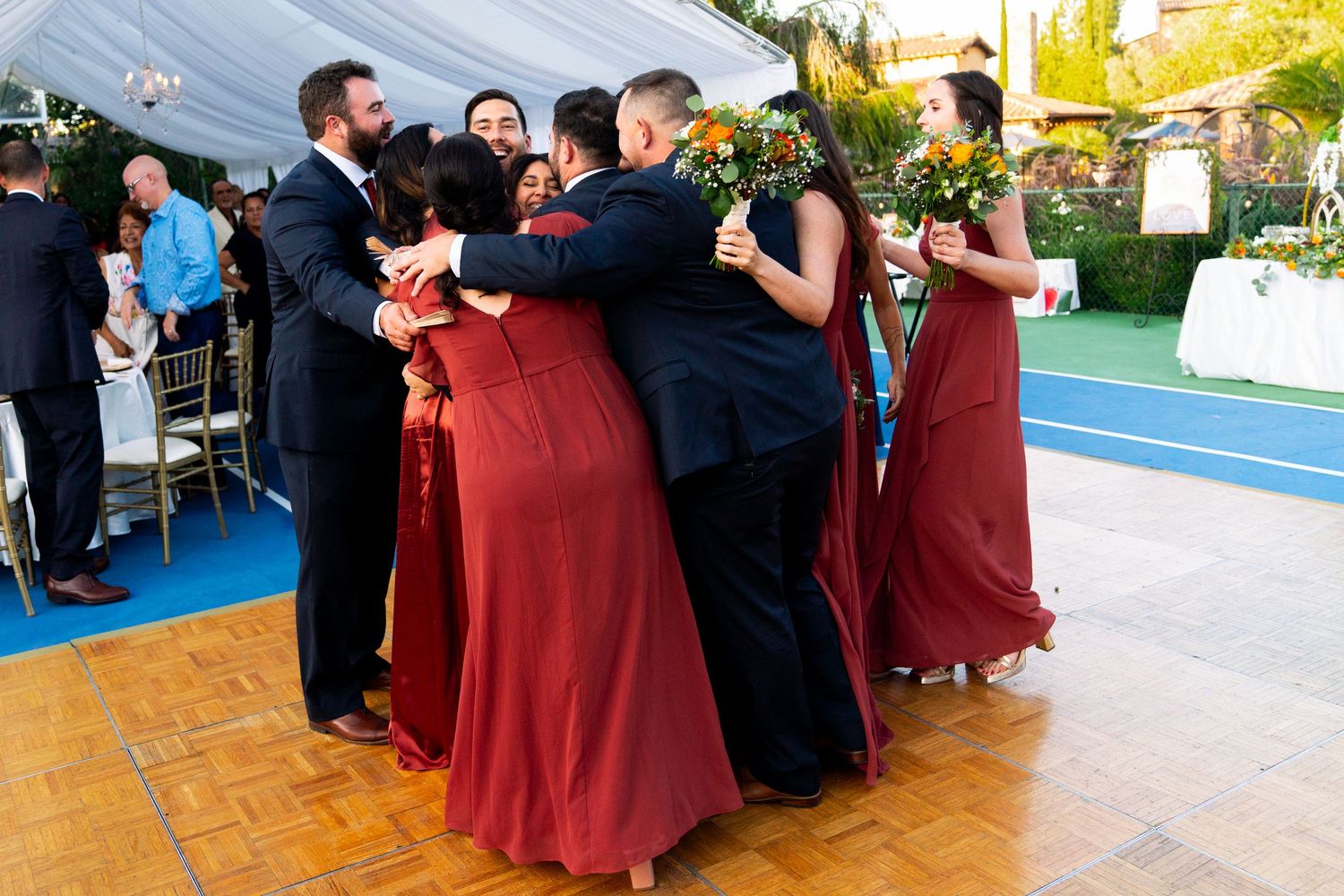 Wedding party dancing together on a blue dance floor under an outdoor tent.