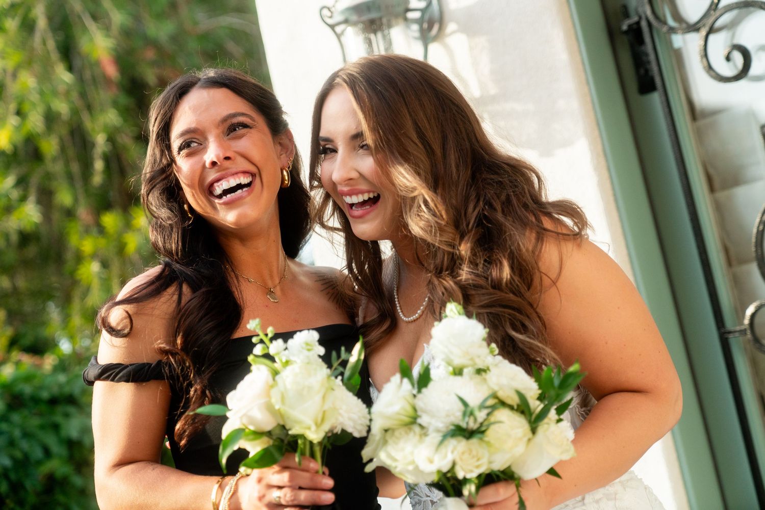 Bridesmaids with wavy dark hair laughing and holding white bouquets at an elegant outdoor venue.