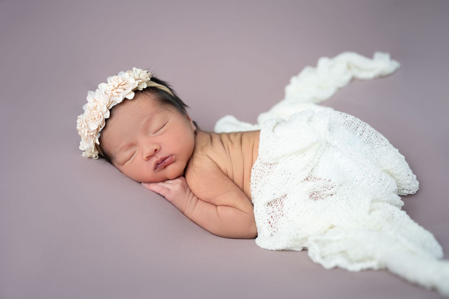 Sweet newborn photo with lace wrap and delicate floral headband against purple backdrop.