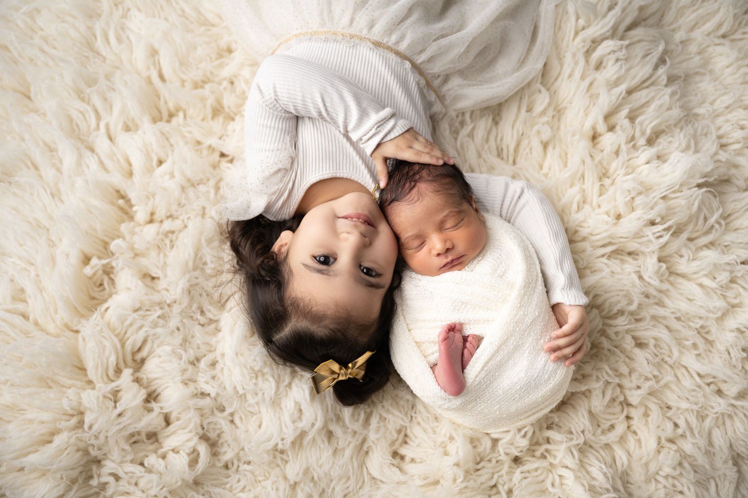 Two children laying together on a cozy cream colored fluffy rug, wearing white clothing.