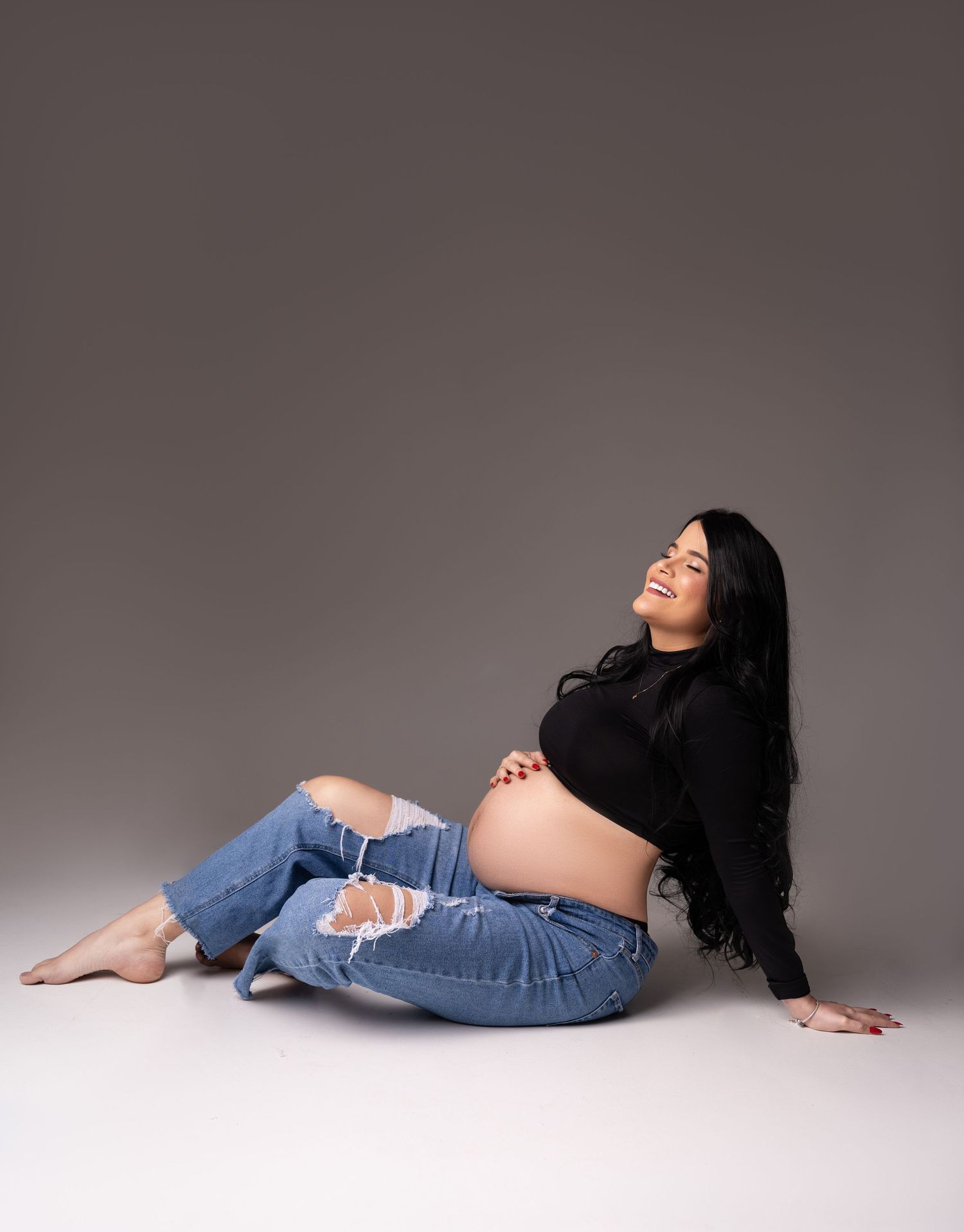 A woman in ripped jeans and a black crop top reclines on the floor during a maternity photo shoot.