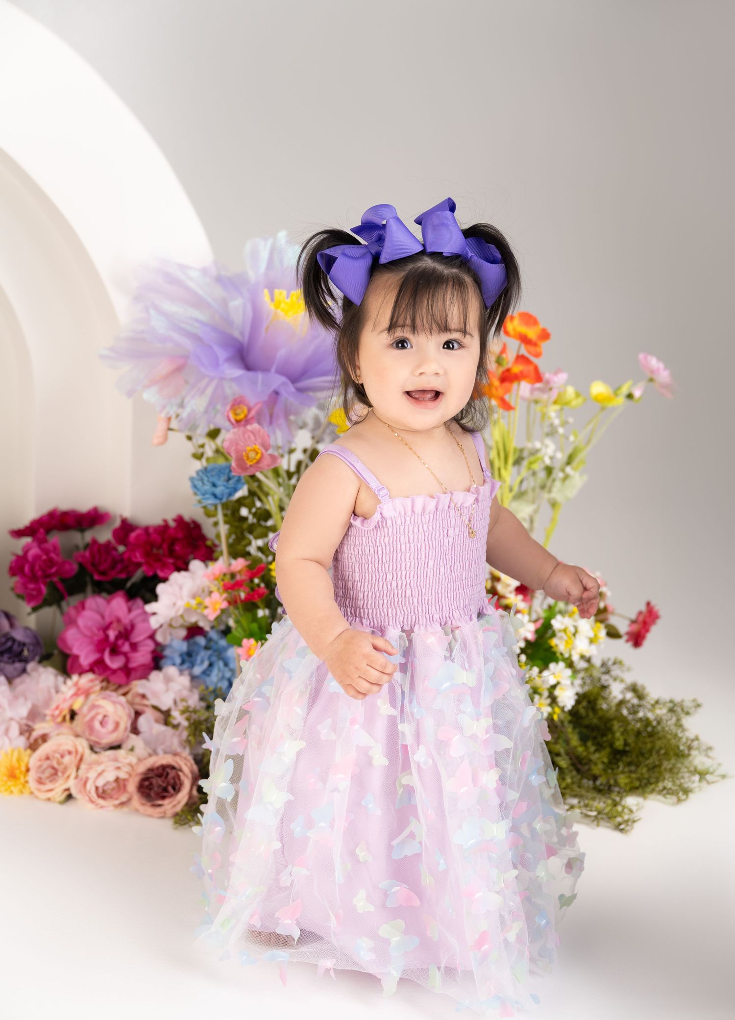 A small child wearing a purple bow and tulle dress poses among colorful flowers.
