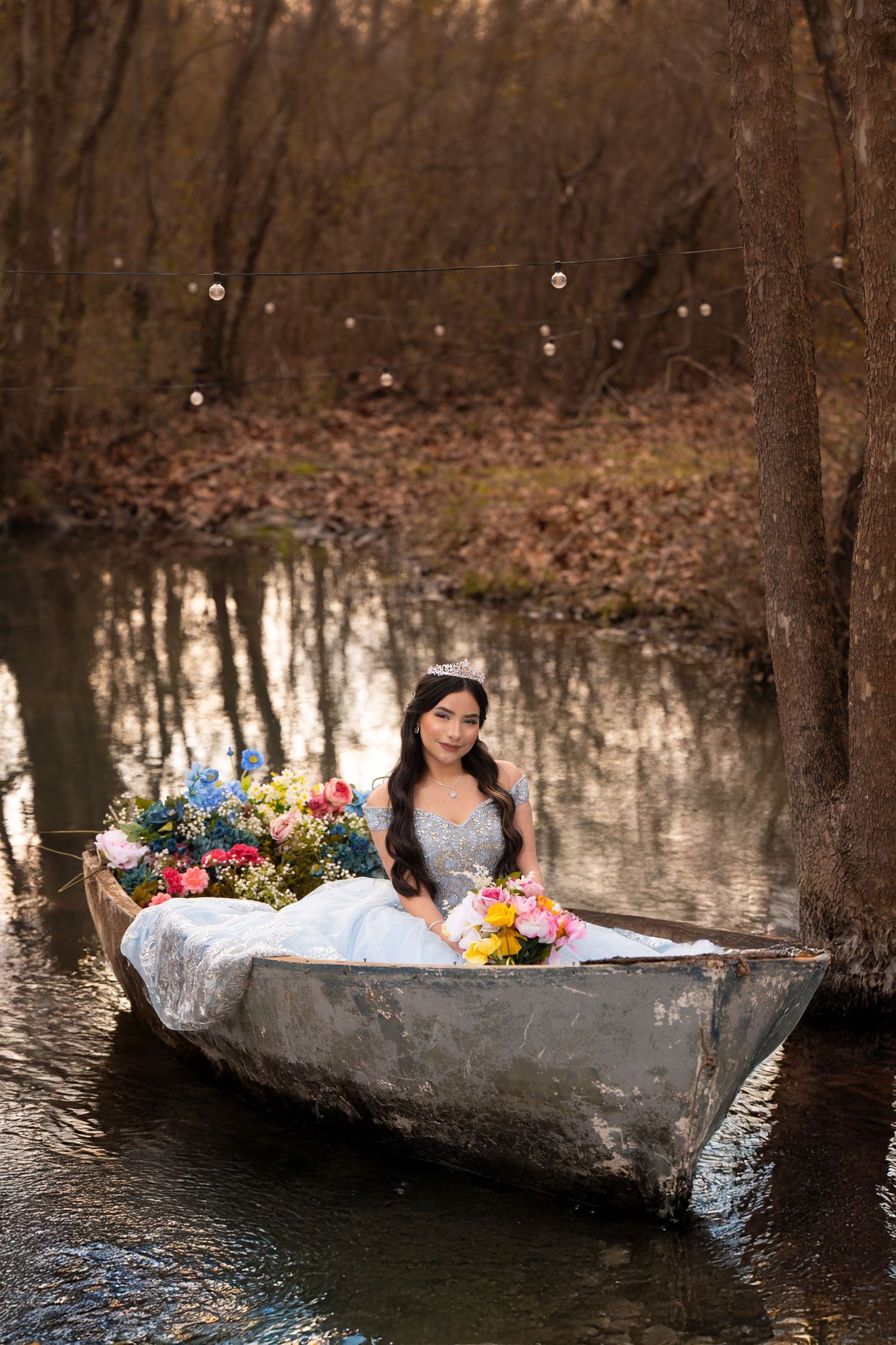 A person in an elegant white dress lies gracefully in a rustic canoe filled with colorful flowers on a tranquil woodland pond.