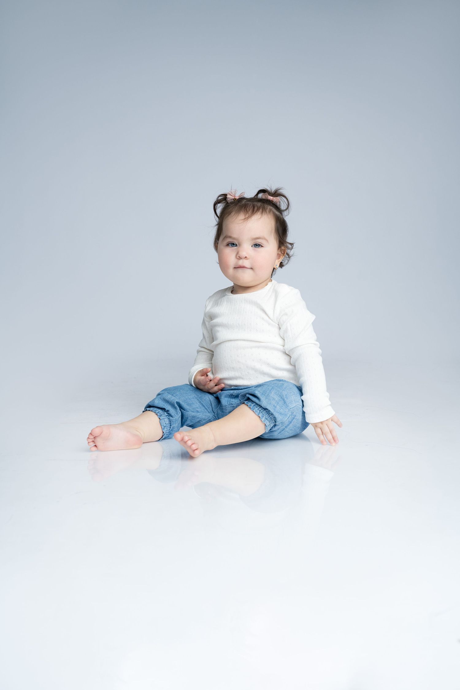 Young toddler in white long sleeve shirt and denim shorts sitting on white background.