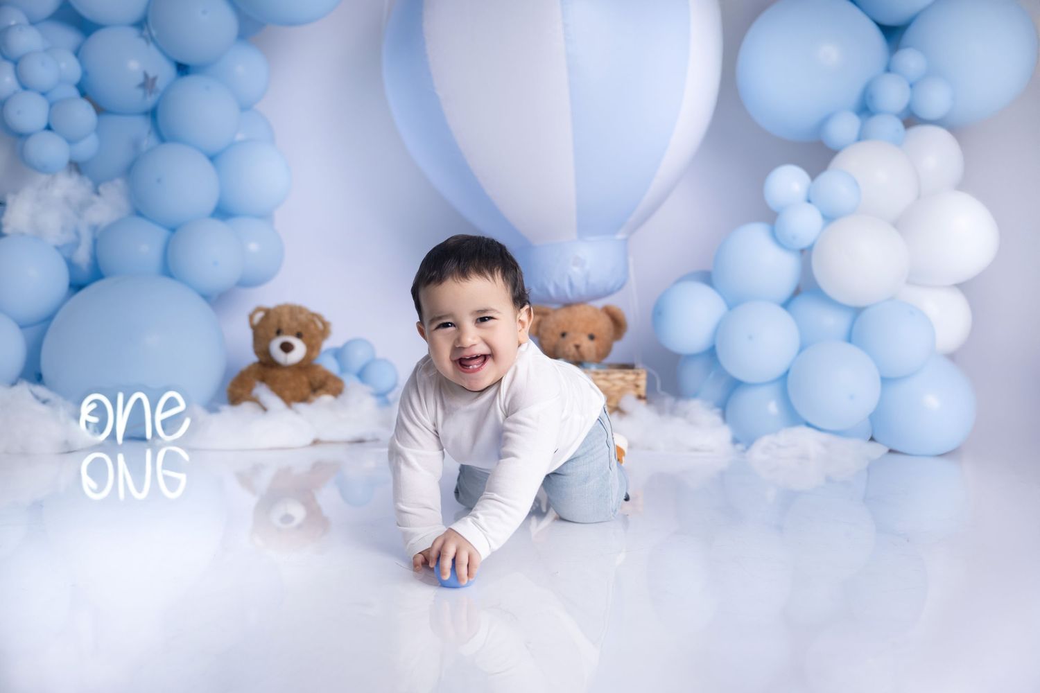 Baby in white outfit crawls among blue and white balloons with teddy bears.