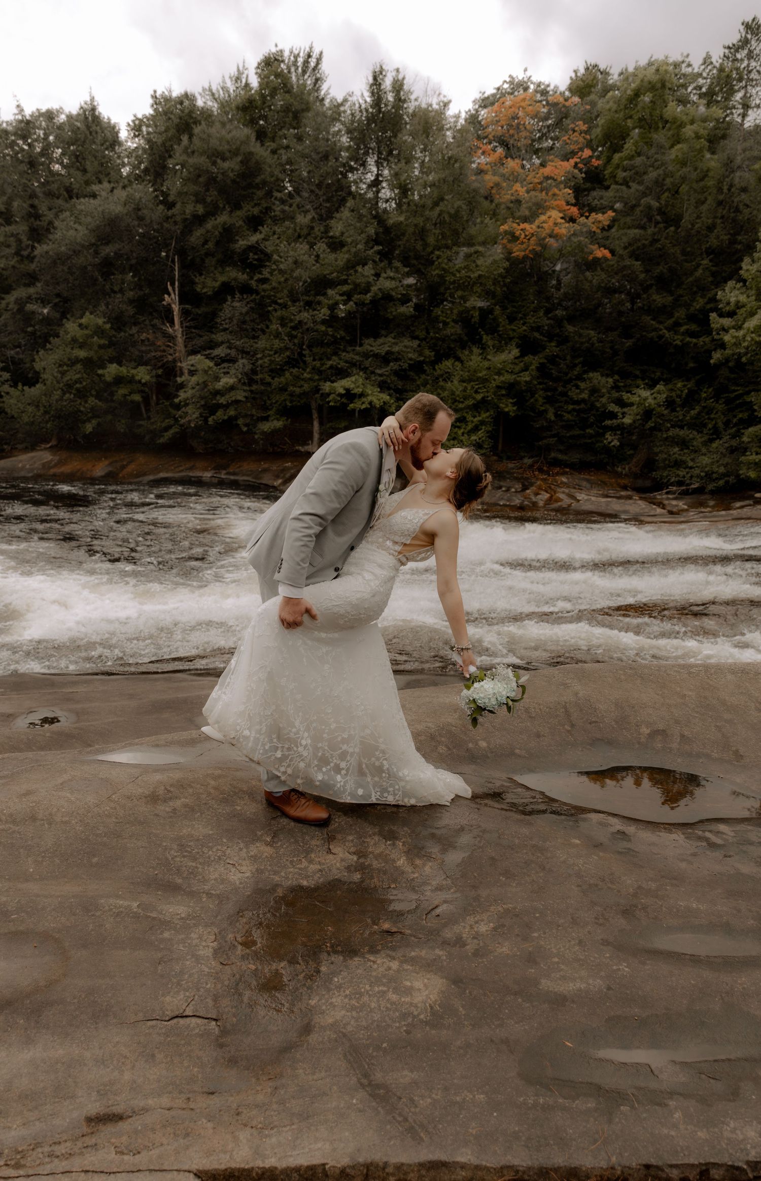 A romantic wedding dip pose on rocks near flowing water with autumn foliage in background.