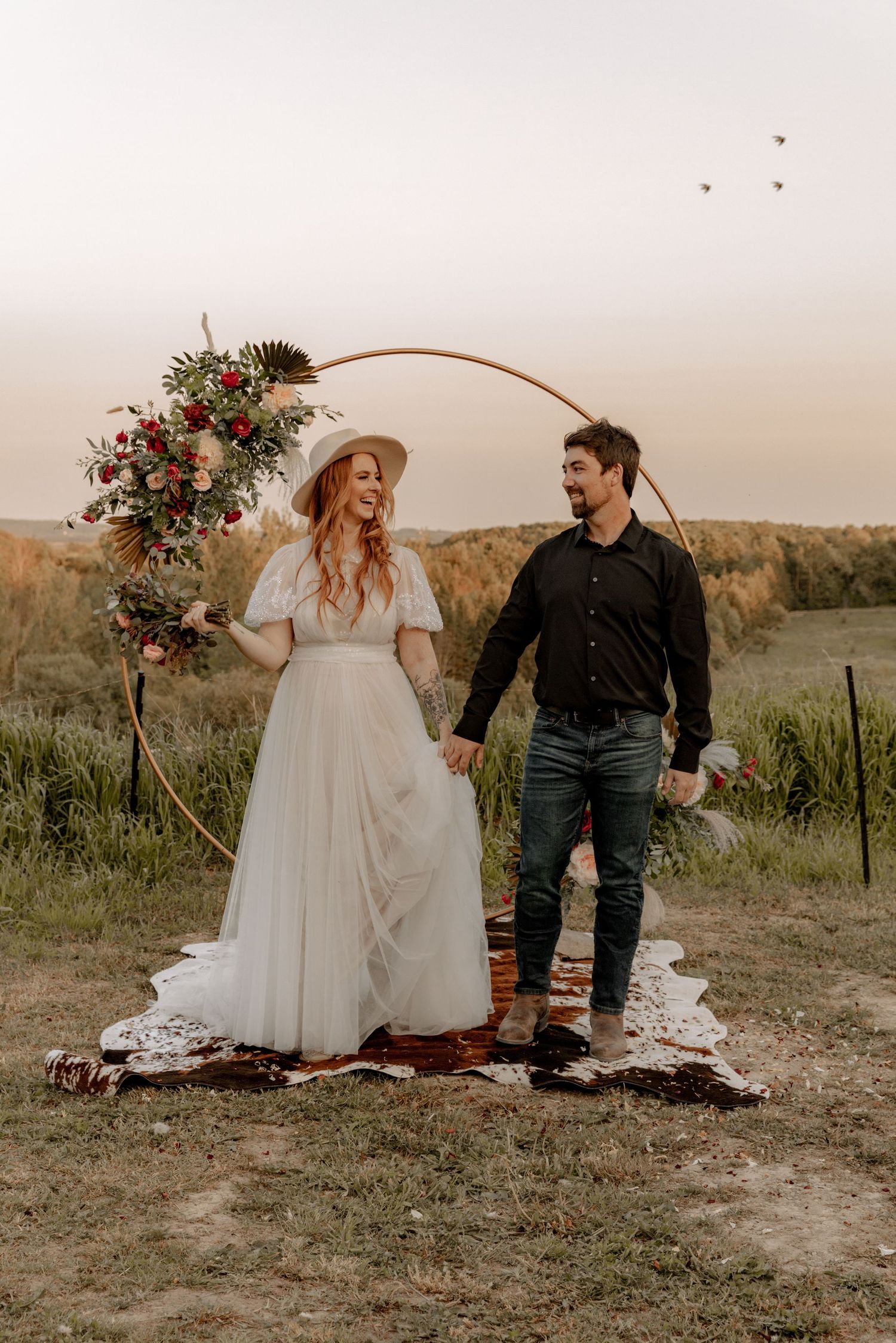 Newlyweds in boho wedding attire pose under curved floral arch at sunset.