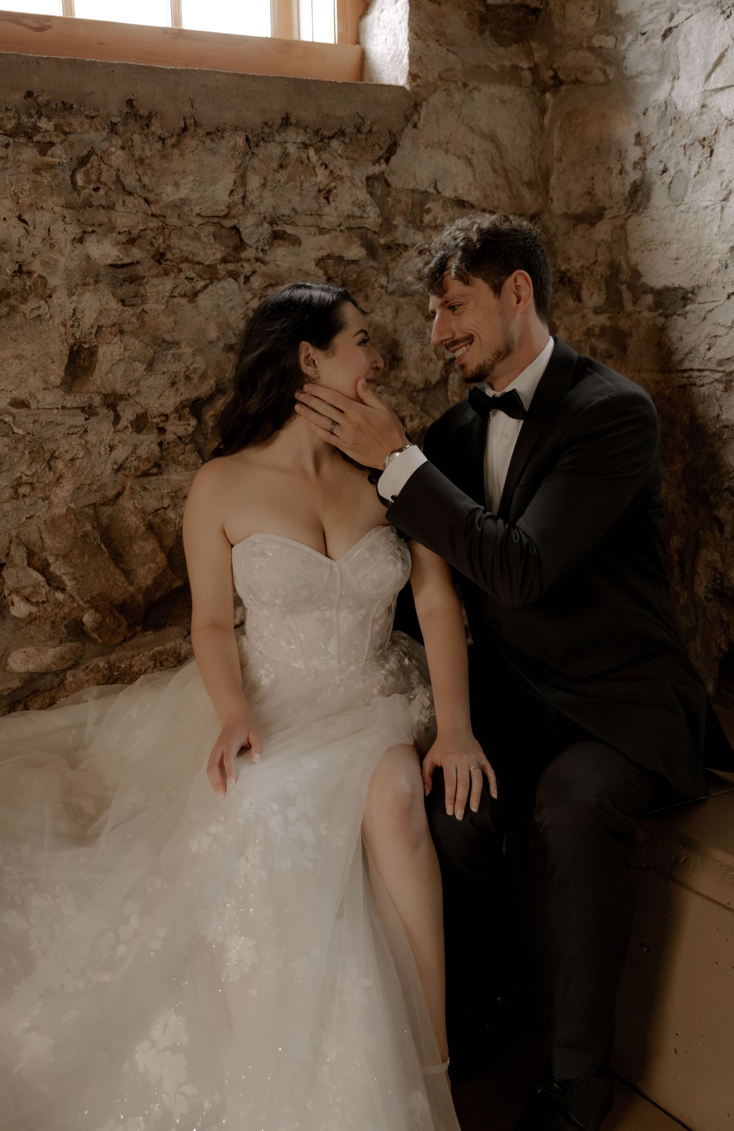 An intimate moment captured between a bride in a white gown and groom in a black suit against rustic stone walls.
