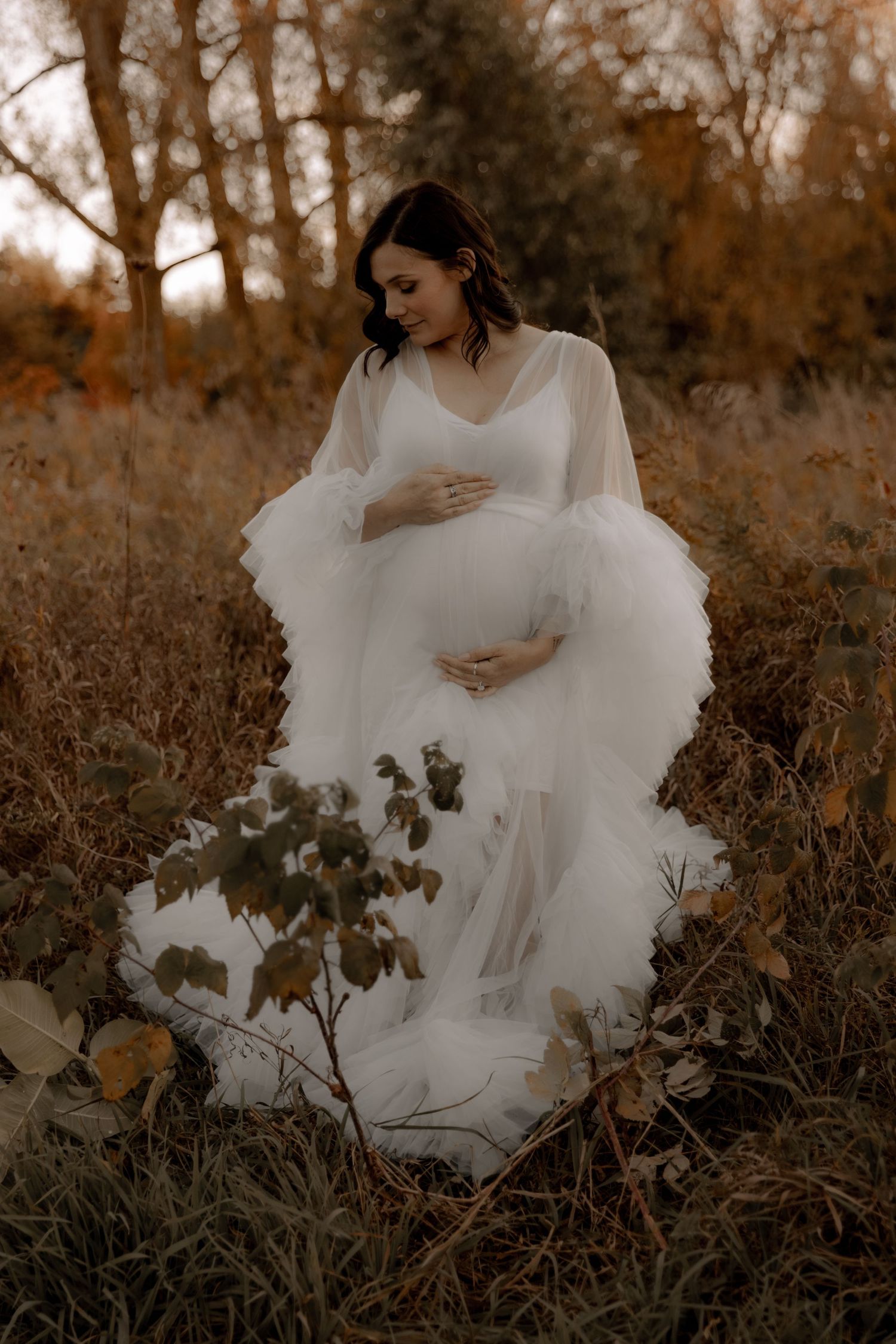 A maternity photoshoot of a flowing white dress in a golden autumn field with tall grass and trees.