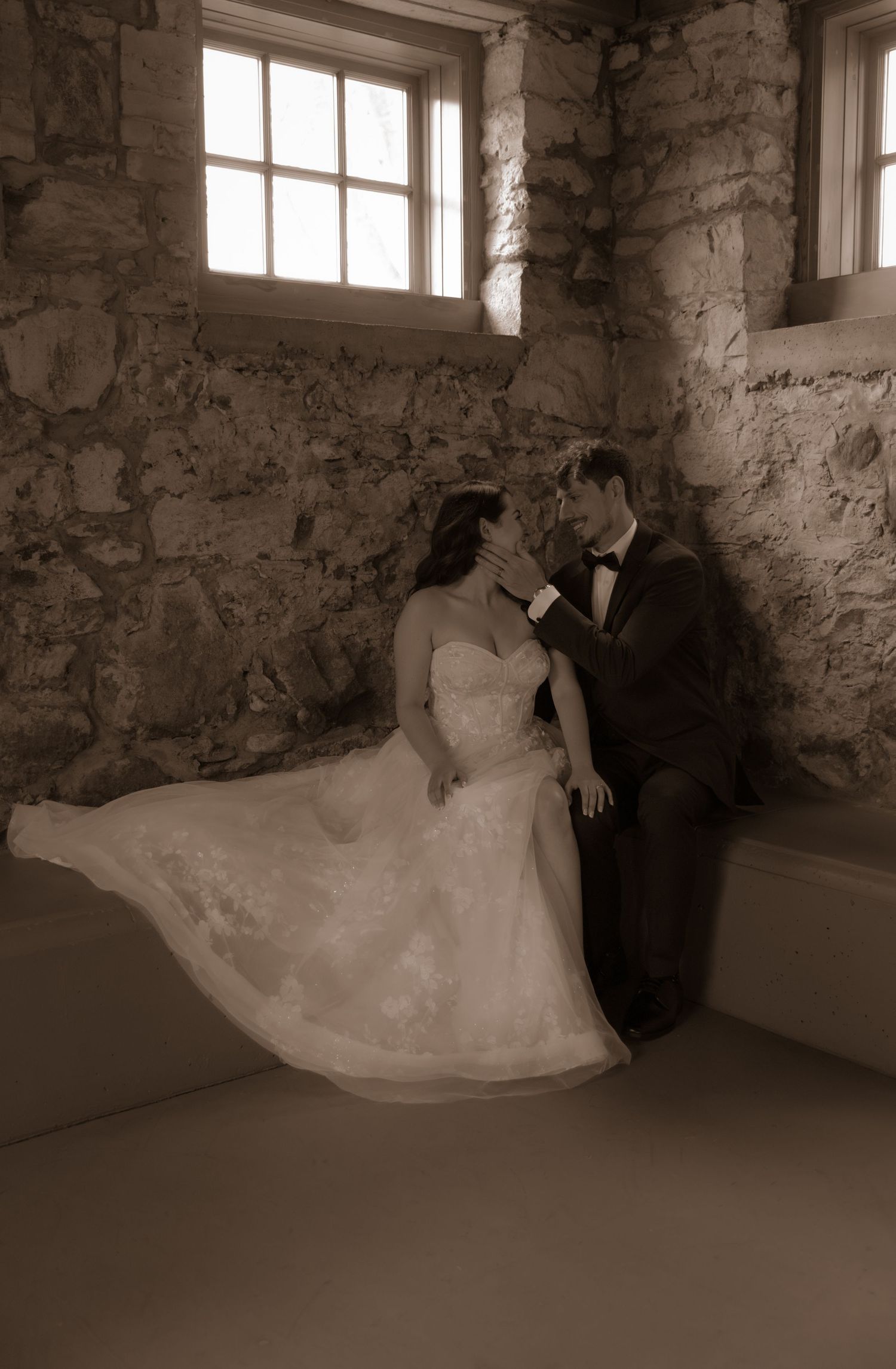 Sepia-toned wedding portrait showing couple in an intimate pose by stone walls and windows.