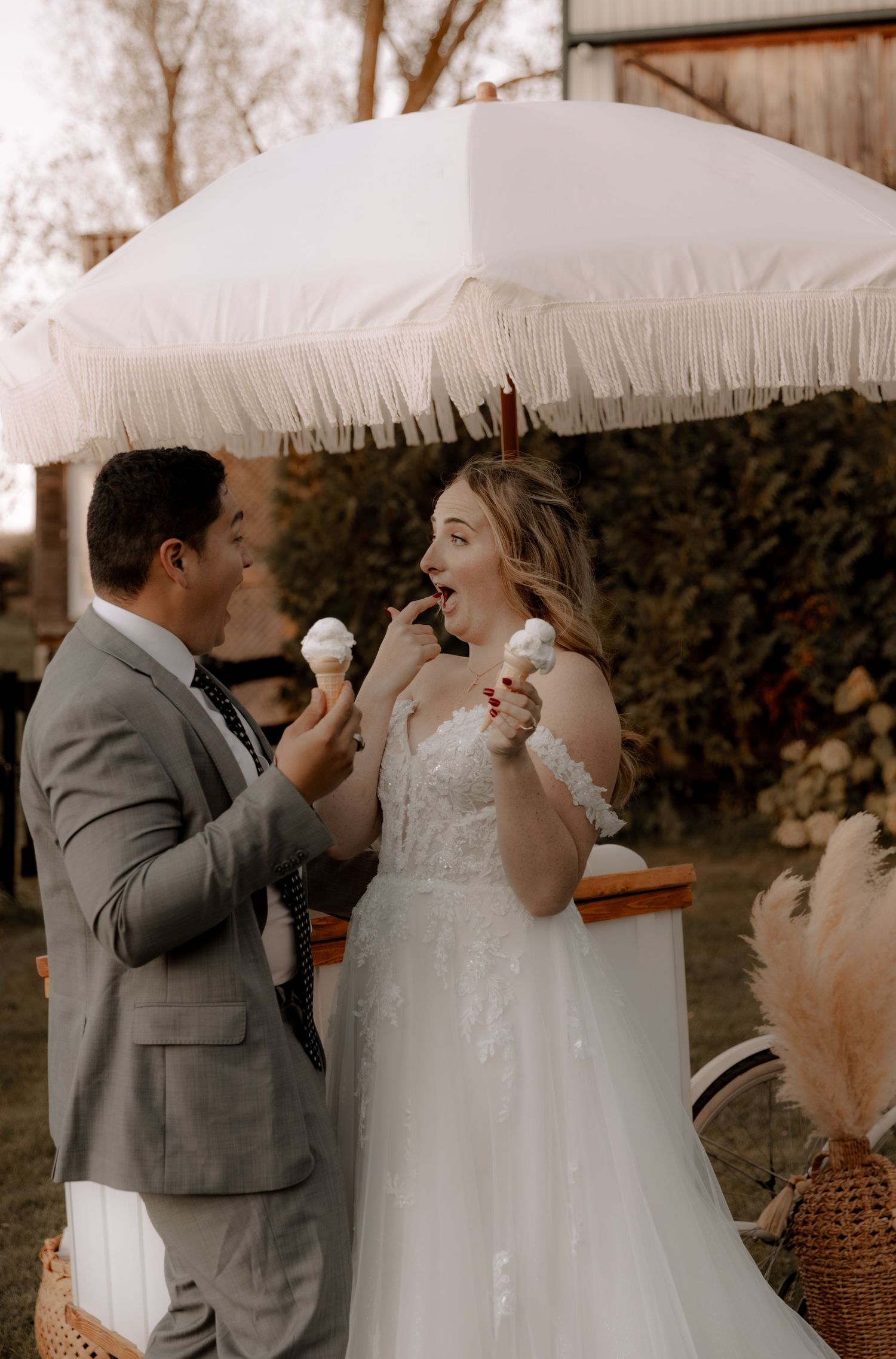 Couple sharing ice cream under white umbrella at rustic outdoor wedding.
