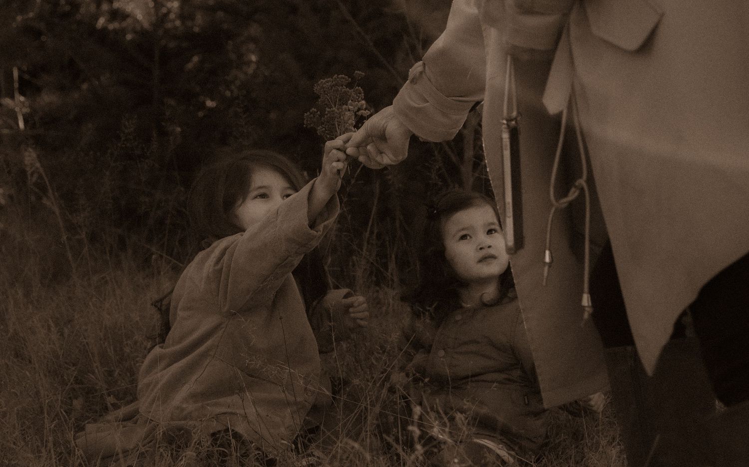 Two small children reaching upward on grass toward an adult's outstretched hand in a sepia-toned photograph.