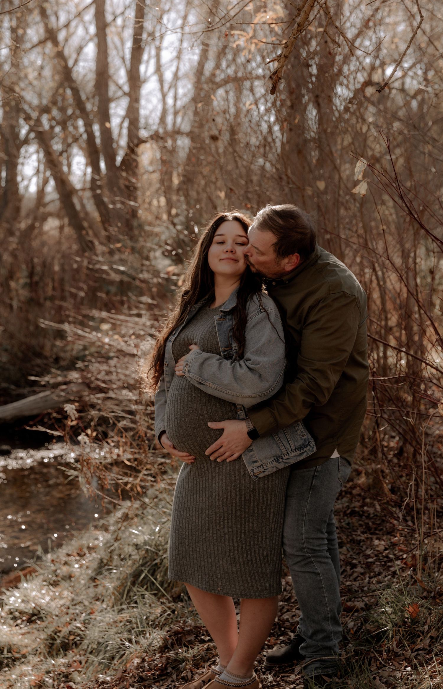 A couple shares an intimate moment in a wooded setting with autumn trees during a maternity photoshoot.