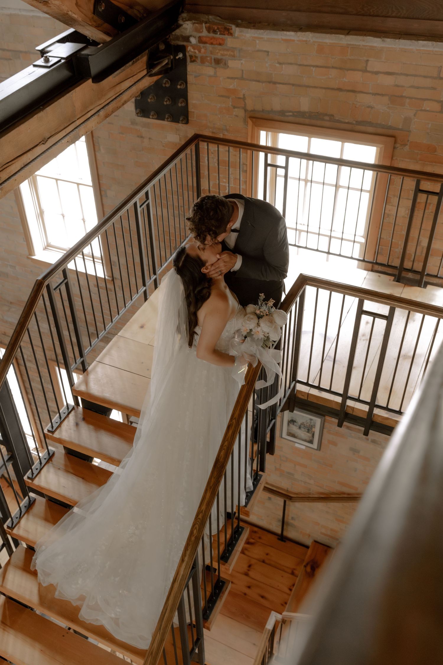 A newlywed couple embracing on a rustic wooden staircase with brick walls and natural lighting.