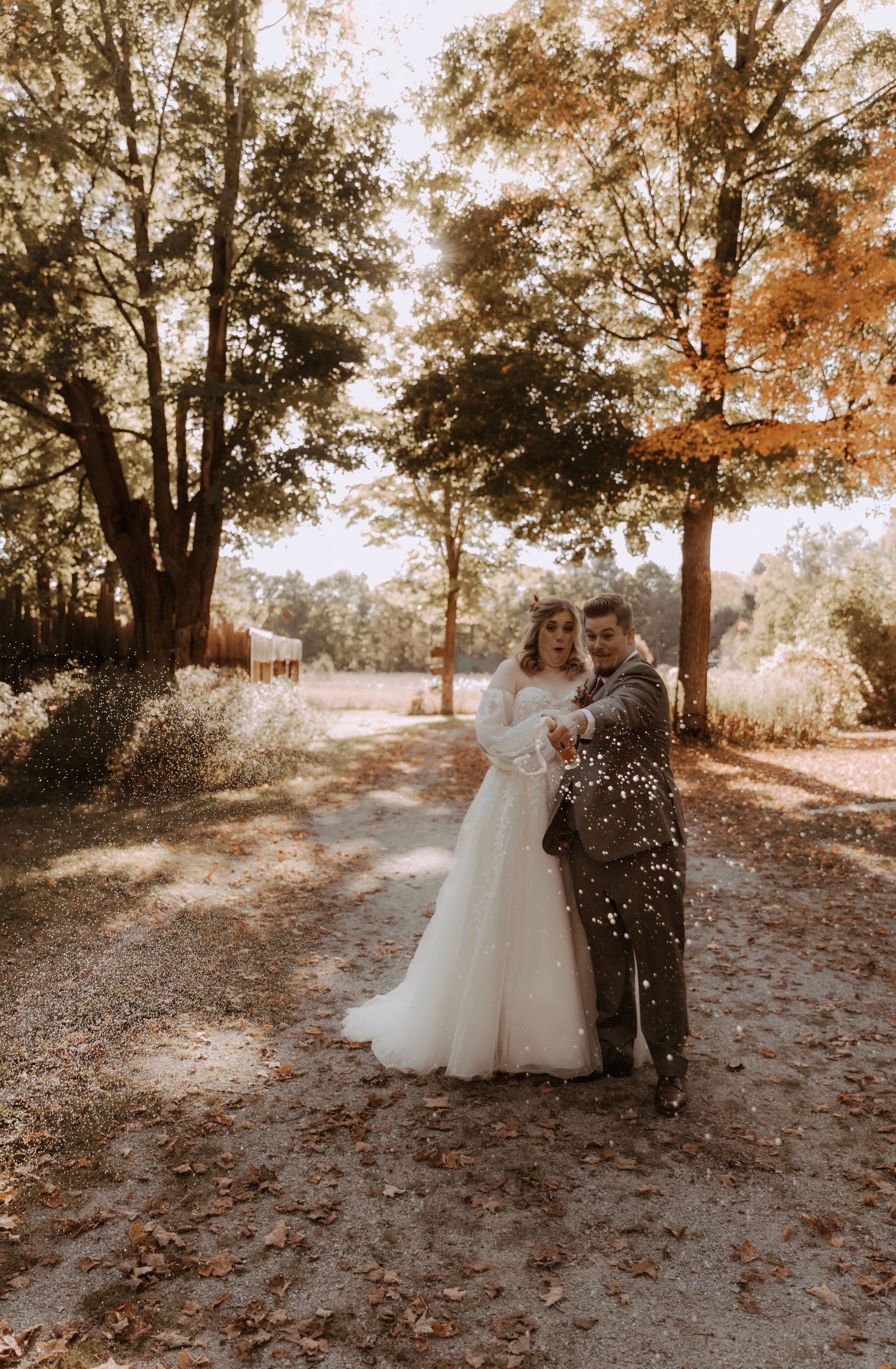 Couple embraces on leaf-covered path with sunlight streaming through autumn trees.
