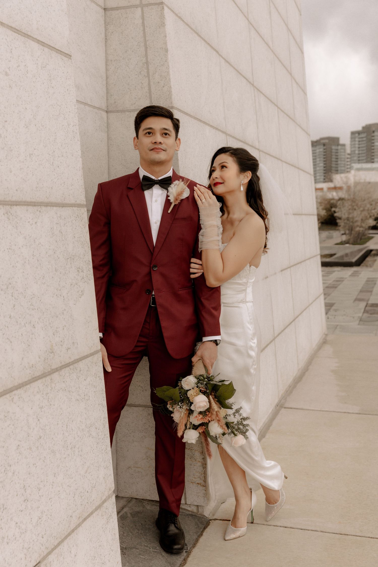 Wedding couple in burgundy suit and white dress pose against modern stone building wall.