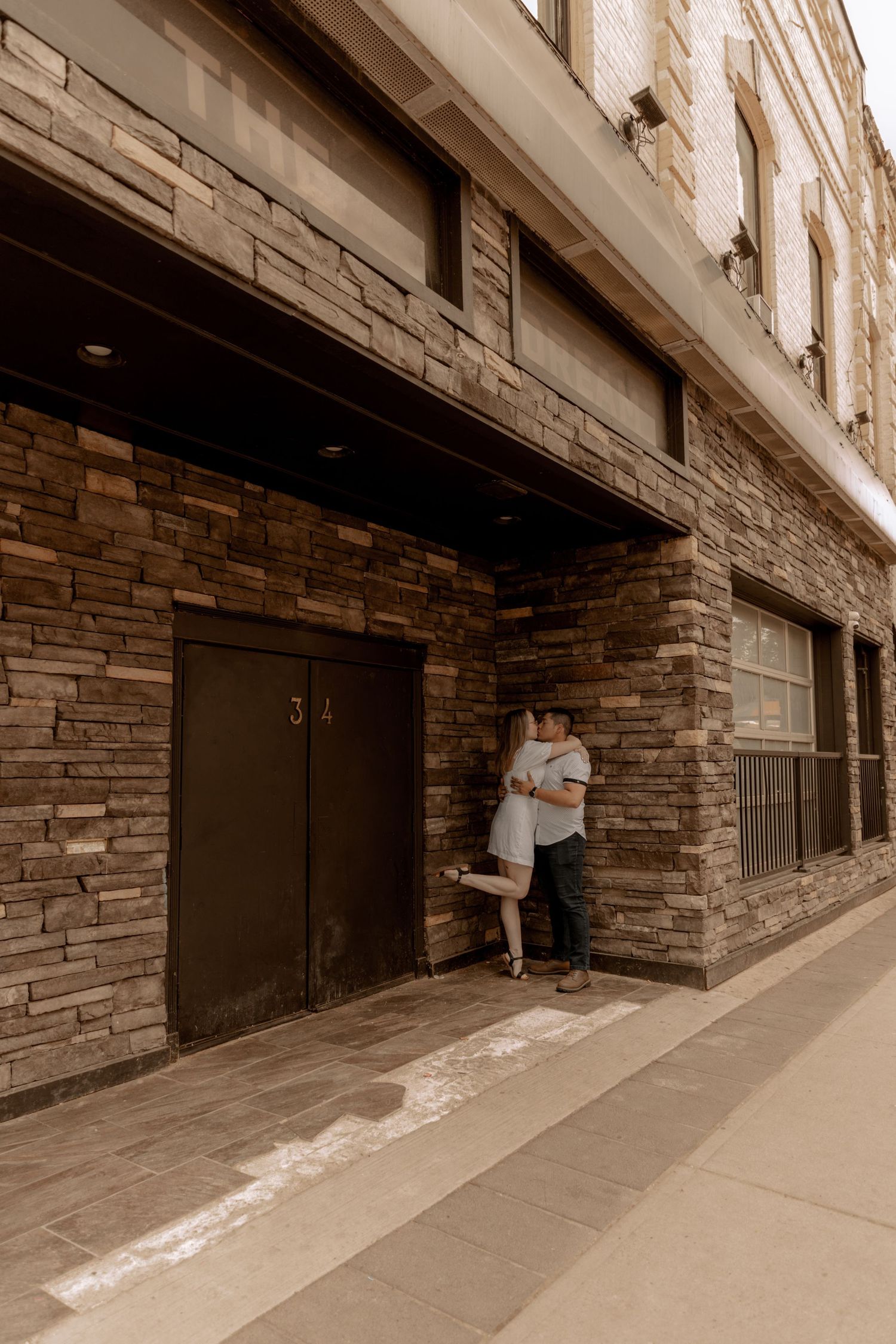 A couple embraces against a rustic stone wall exterior of an urban building in sepia tones.