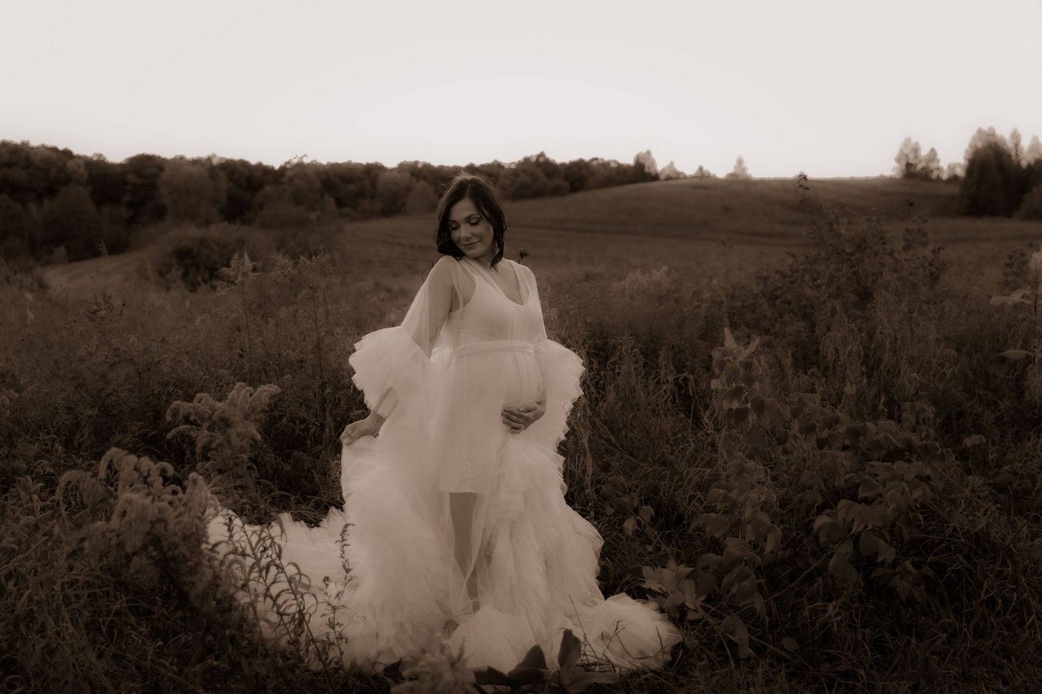 A graceful figure in a flowing white dress poses against a serene rural landscape at sunset in sepia tones.