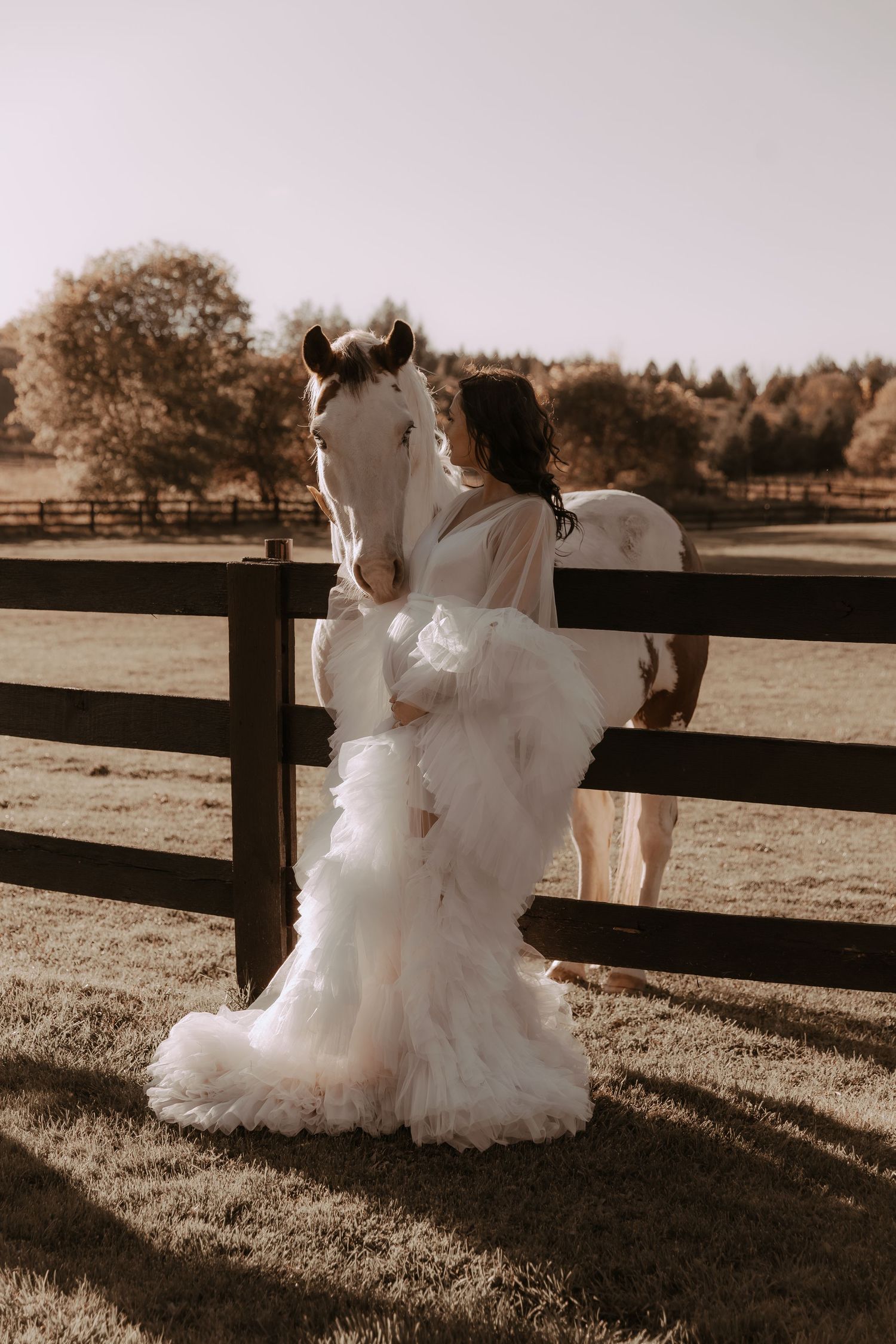 A white horse and flowing wedding dress create a romantic scene against a rustic fence in warm sepia tones.